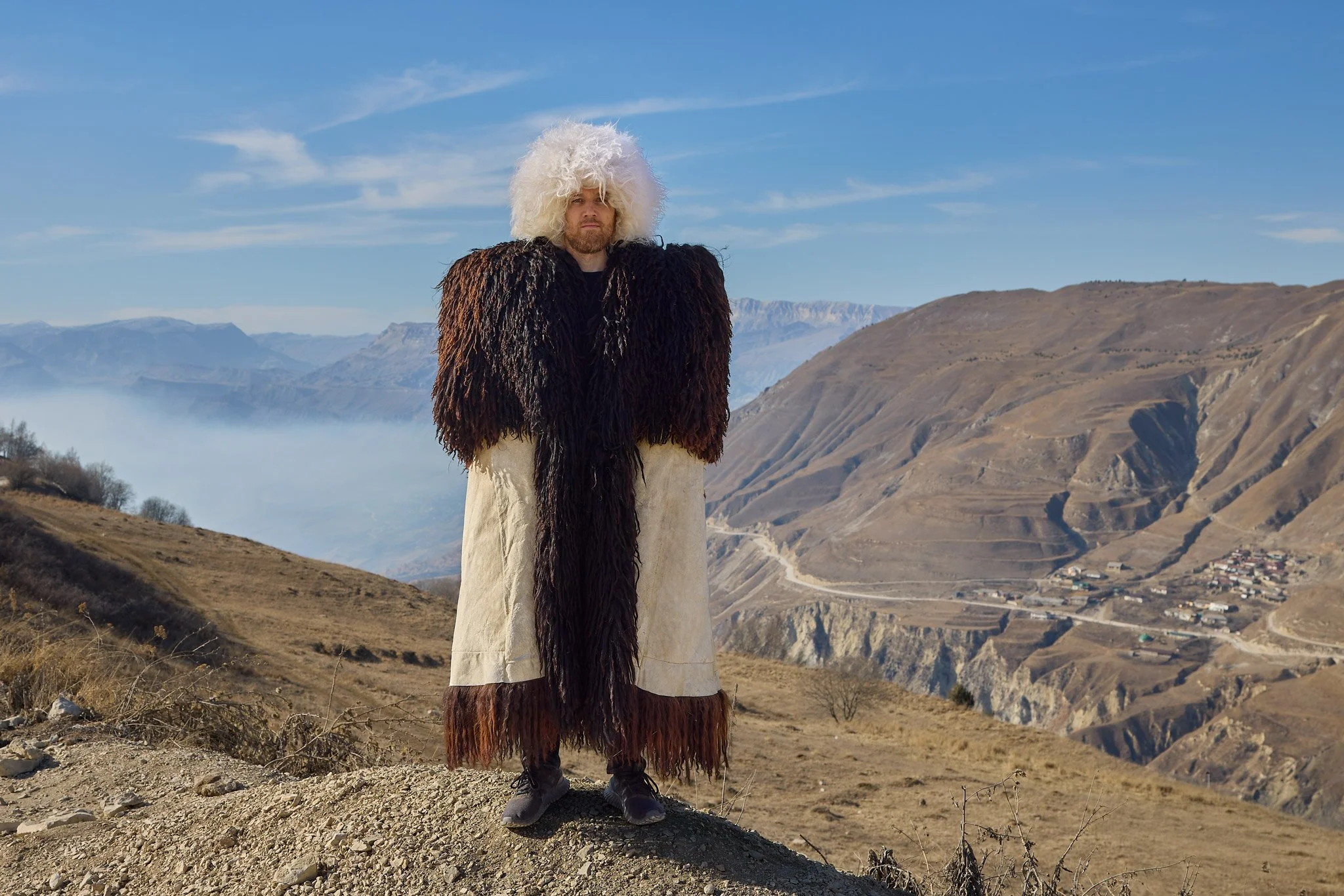 A man standing outdoors in a mountainous landscape, wearing a large leather coat with shaggy fur details and black shoes, with mountains and a small village in the background.