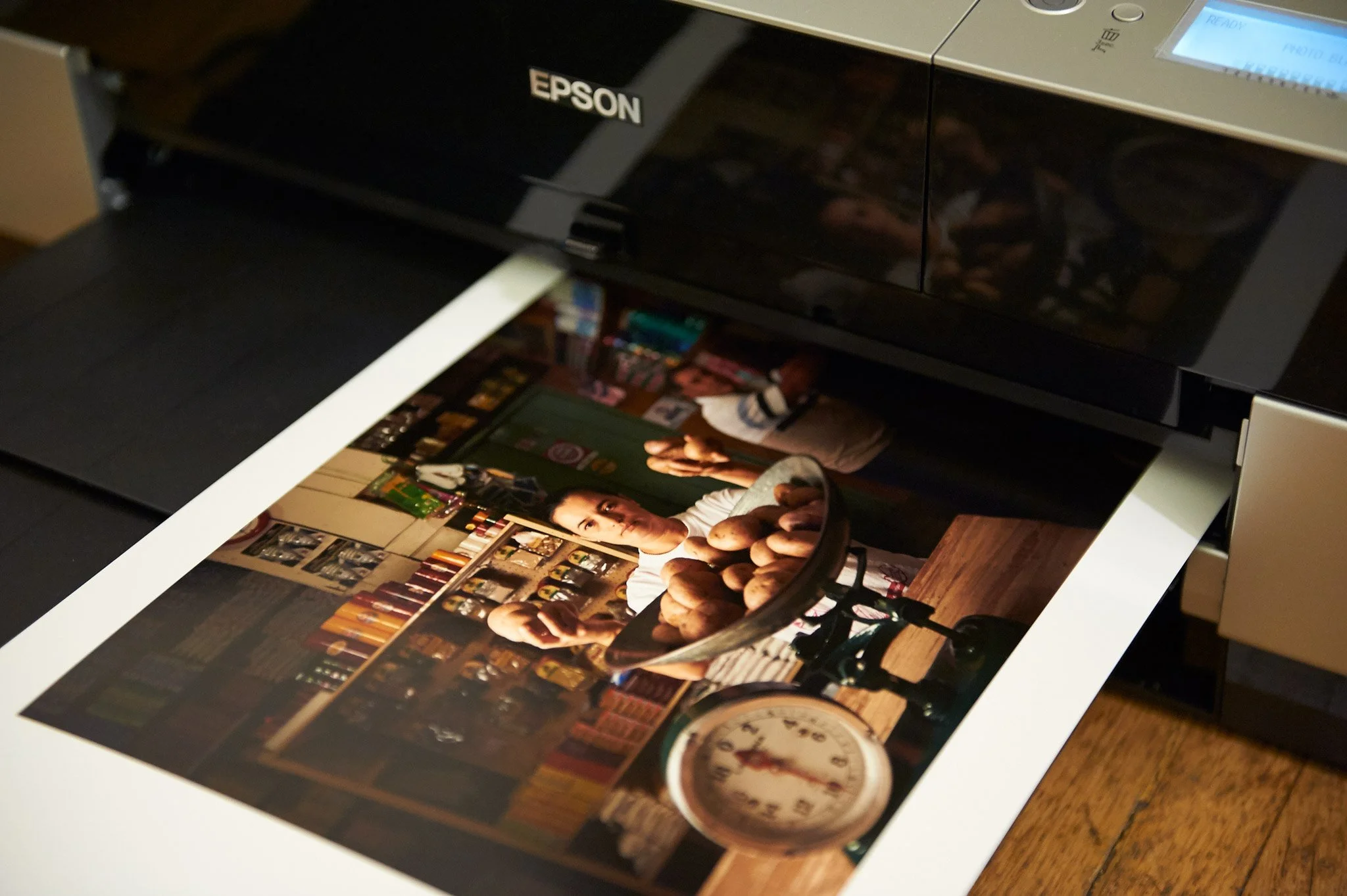 Photograph of a woman in a store, reaching towards the camera with a spoon, with a scale and potatoes on a table, reflected in a large printer.