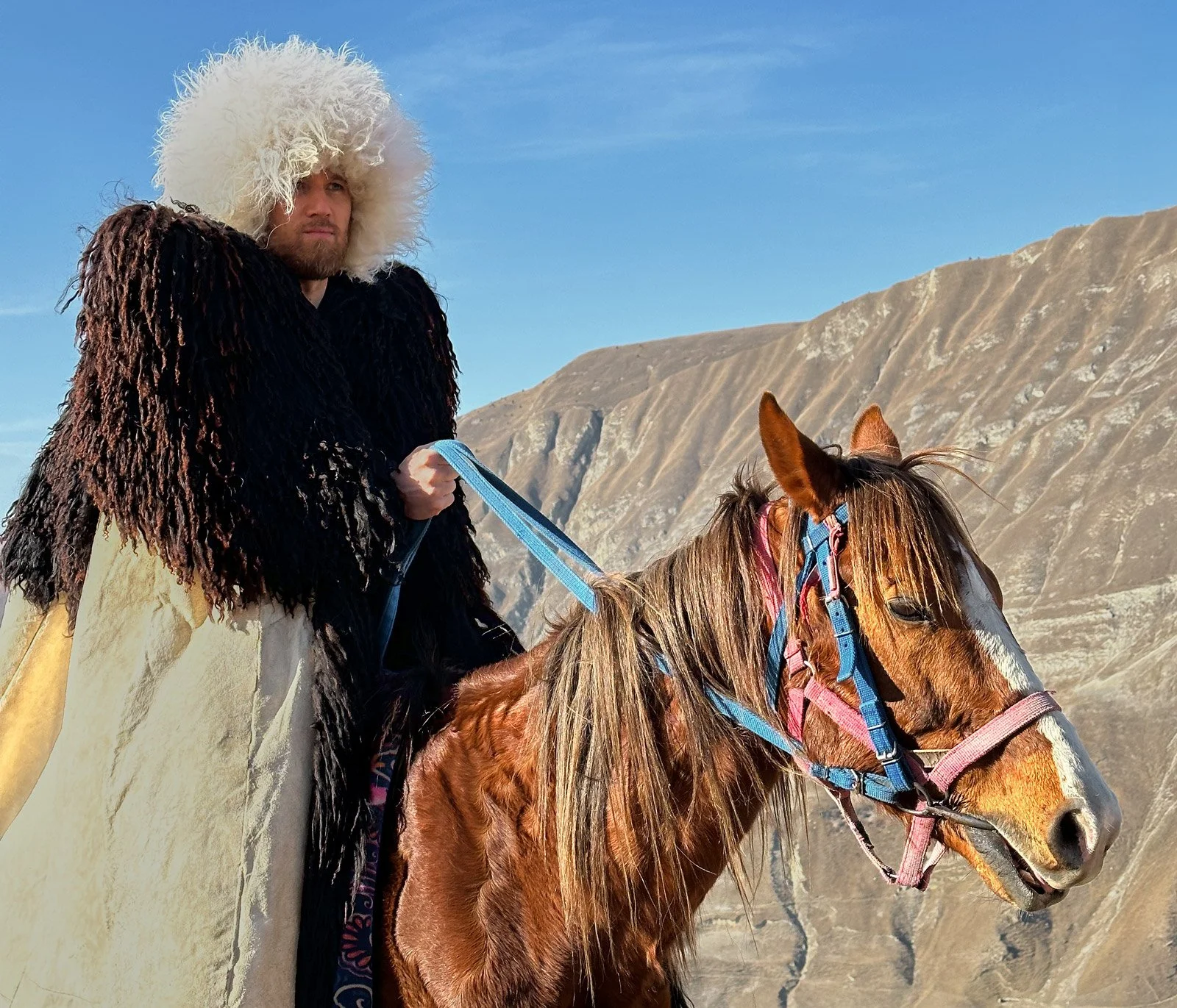 A person dressed in traditional mountaineer attire, with a large white curly hat and dark fur coat, riding a chestnut horse in a mountainous landscape with dry hills and a blue sky.