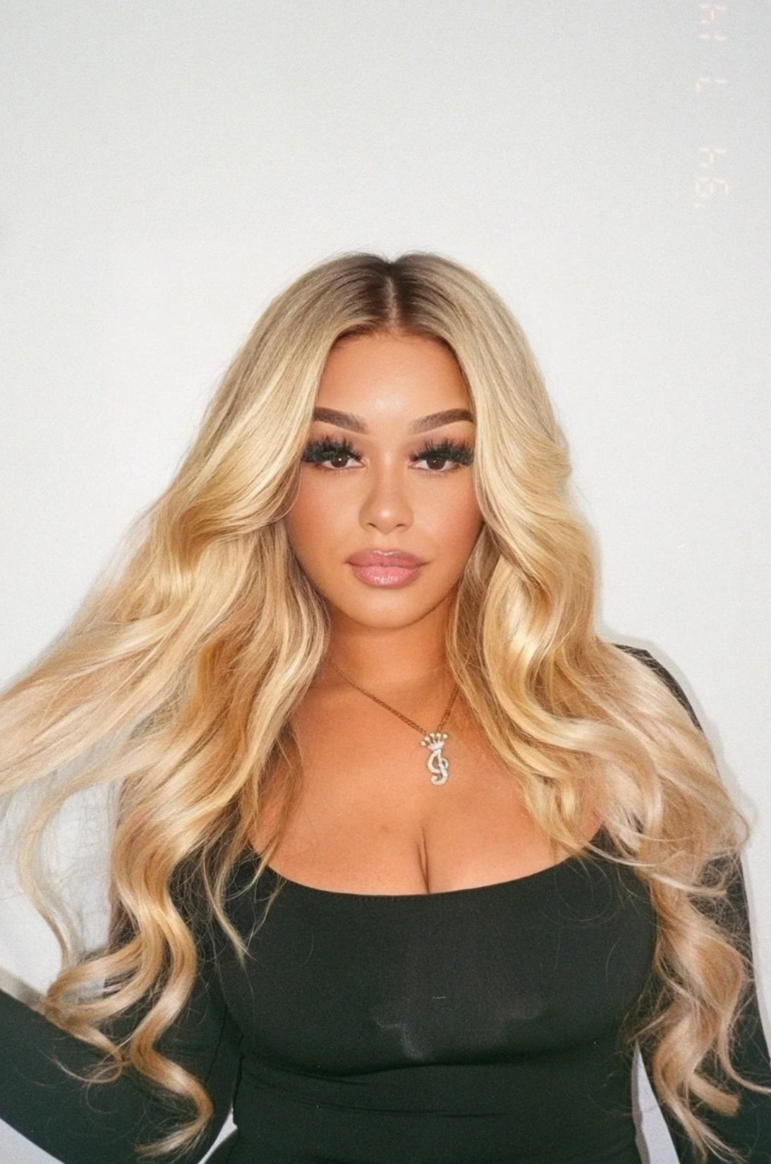 A woman with long, wavy blonde hair and makeup, wearing a black top and a necklace with a pendant, standing against a plain white background.