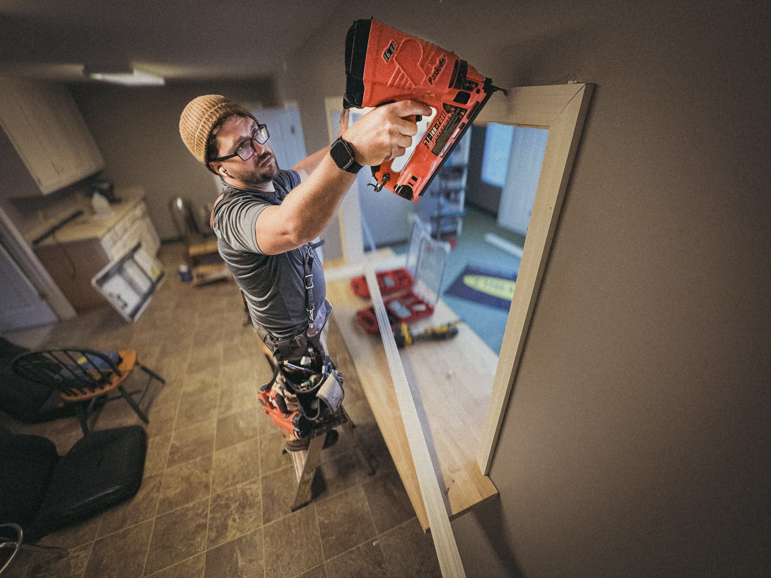 Handyman standing on a short ladder, installing window trim to a service window with an orange paslode finisher nailer