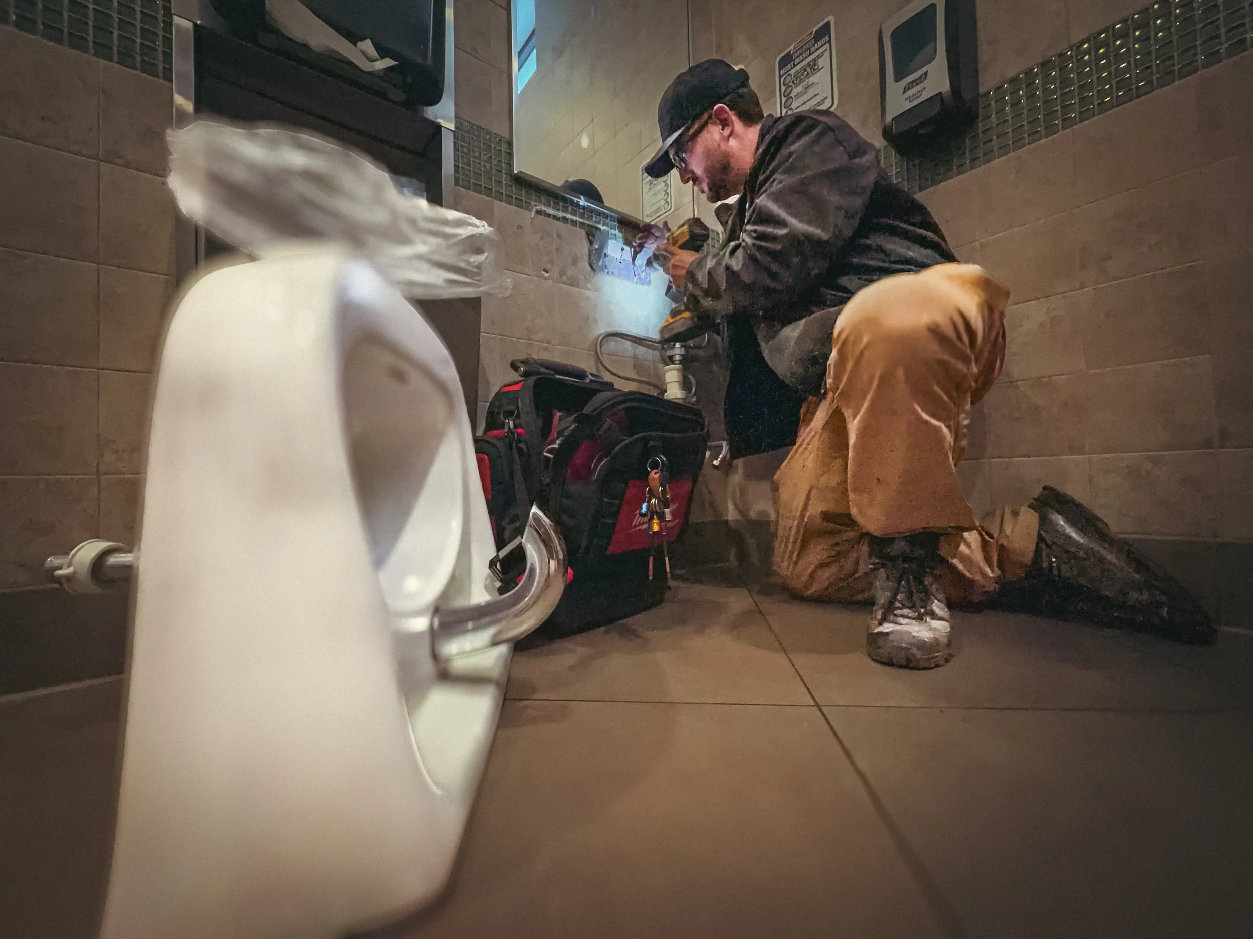 Commercial handyman in an East Atlanta convenience store bathroom, reattaching a commercial sink mount to the wall, with the sink laying nearby. Drill in hand, and technician bag by his side.