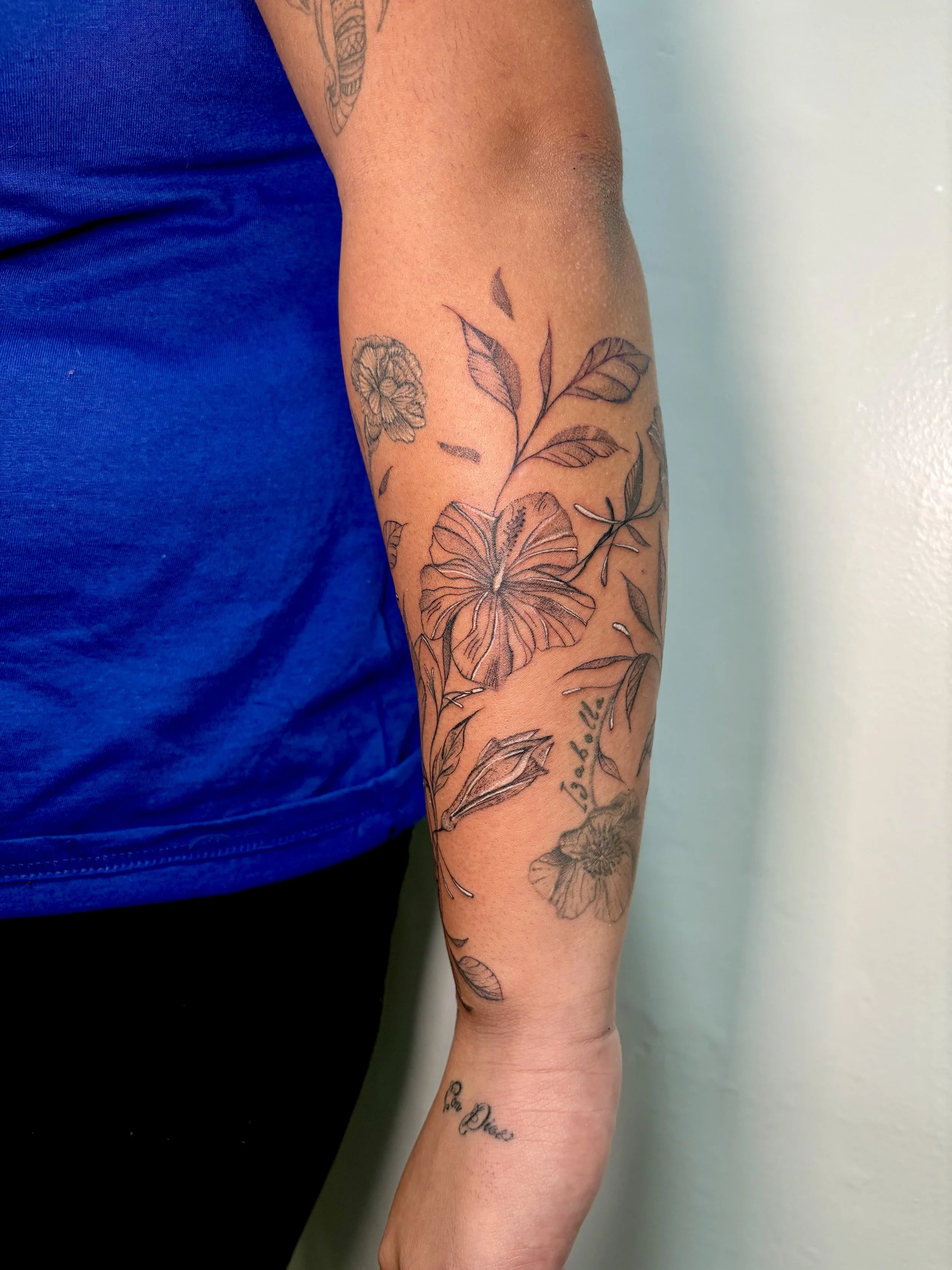 A person's arm with a floral tattoo sleeve, featuring leaves, flowers, and a handwritten script, against a light-colored wall background.