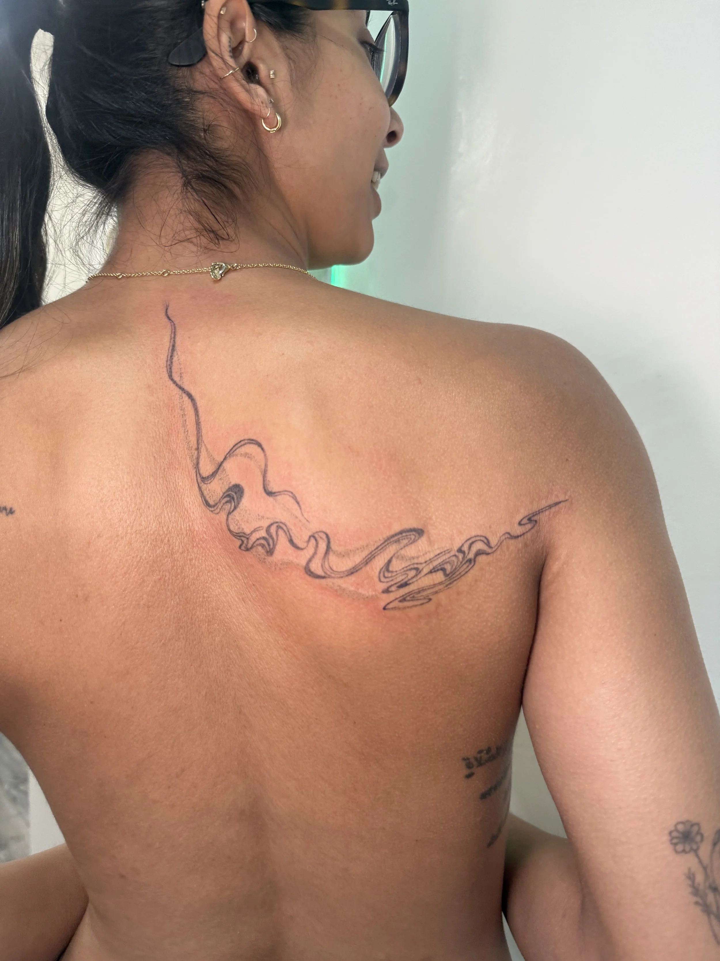 A woman with dark hair in a ponytail, glasses, and multiple earrings, is turned to the side showing her shoulder and back, with a black and red tattoo of smoke or abstract lines on her shoulder.