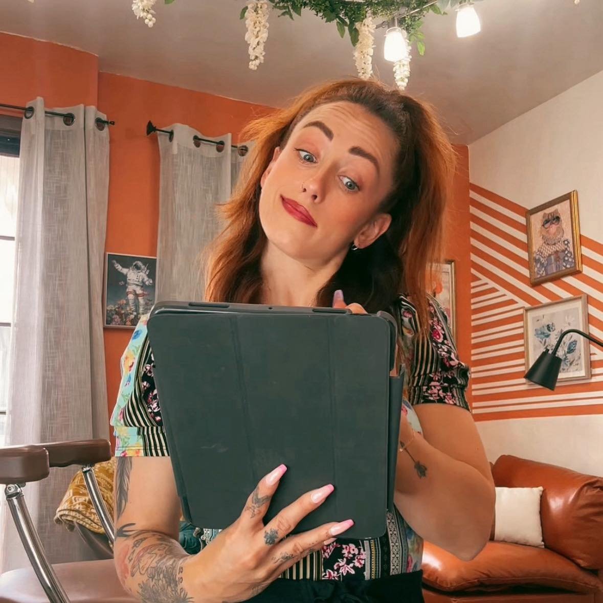 A woman with red hair, tattoos on her arms, and red lipstick looking at a tablet with a skeptical expression in a cozy living room with orange and white walls, framed art, curtains, and decorative ceiling hanging plants.