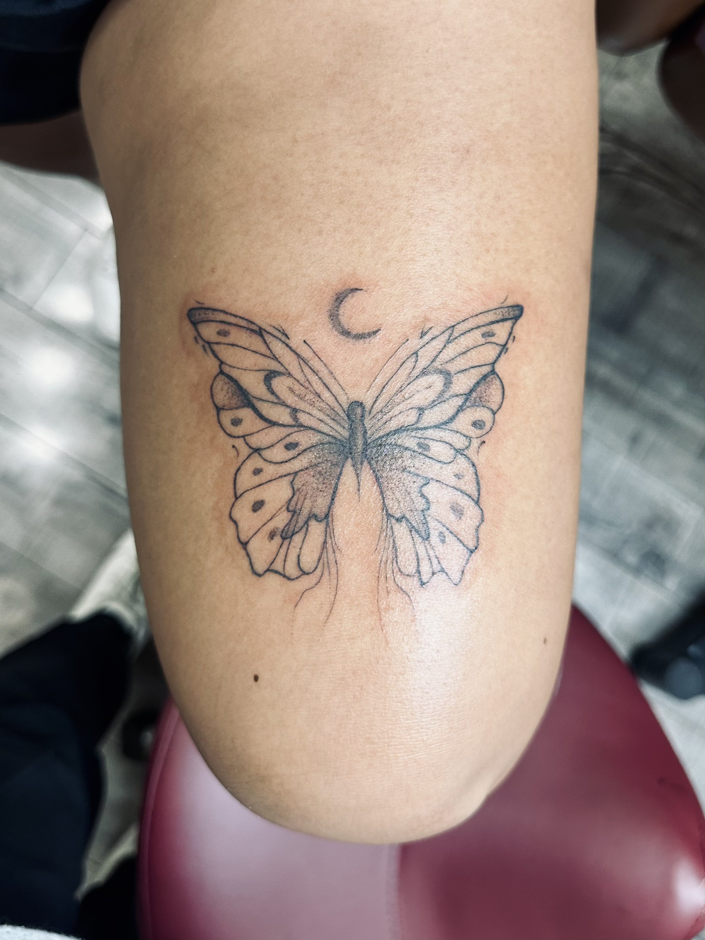 A tattoo of a butterfly with detailed wings above a small crescent moon on a person's thigh.