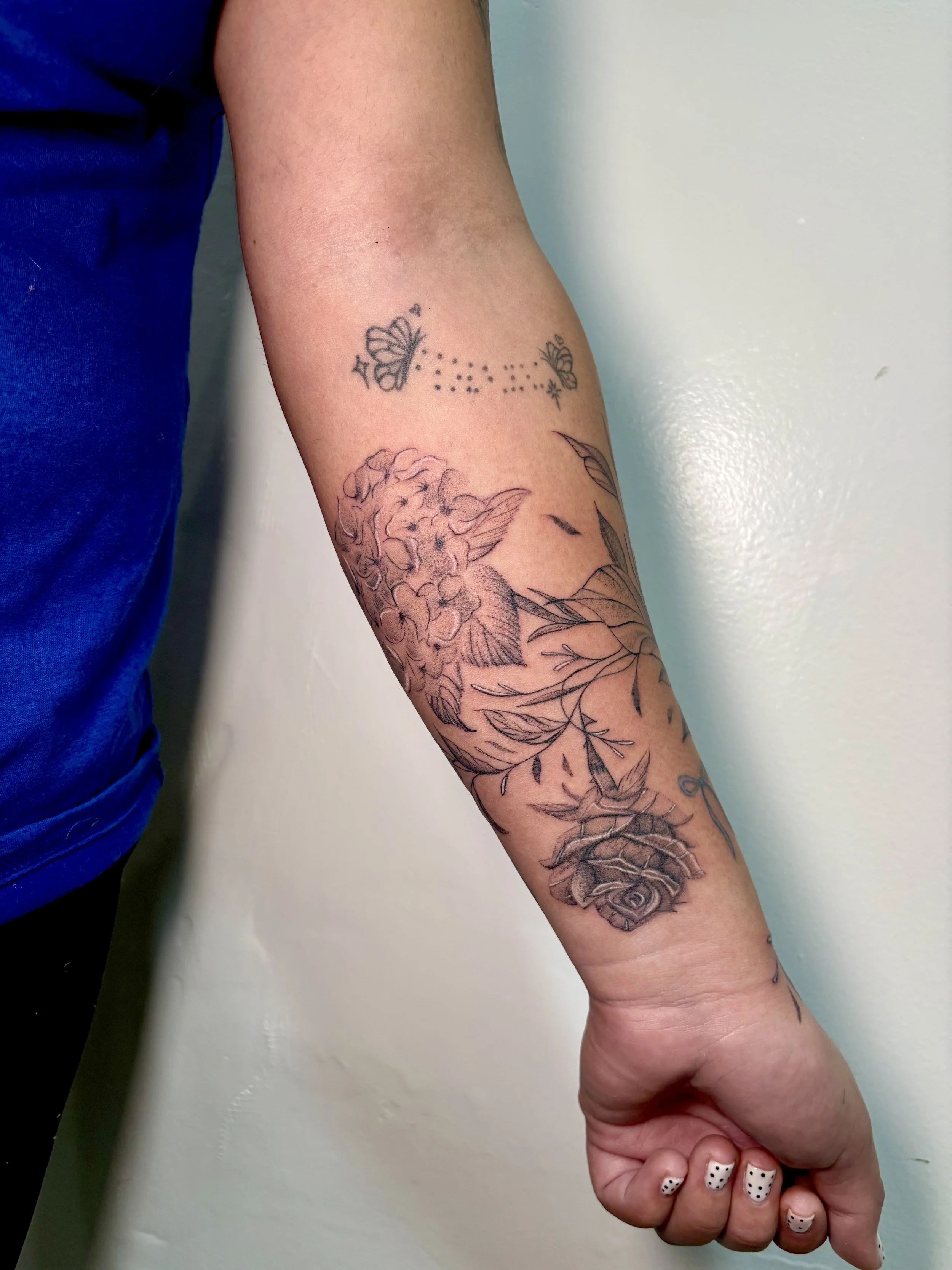 A person's arm with a tattoo of a large flower and leaves, plus a small constellation tattoo of a butterfly with stars and small dots.