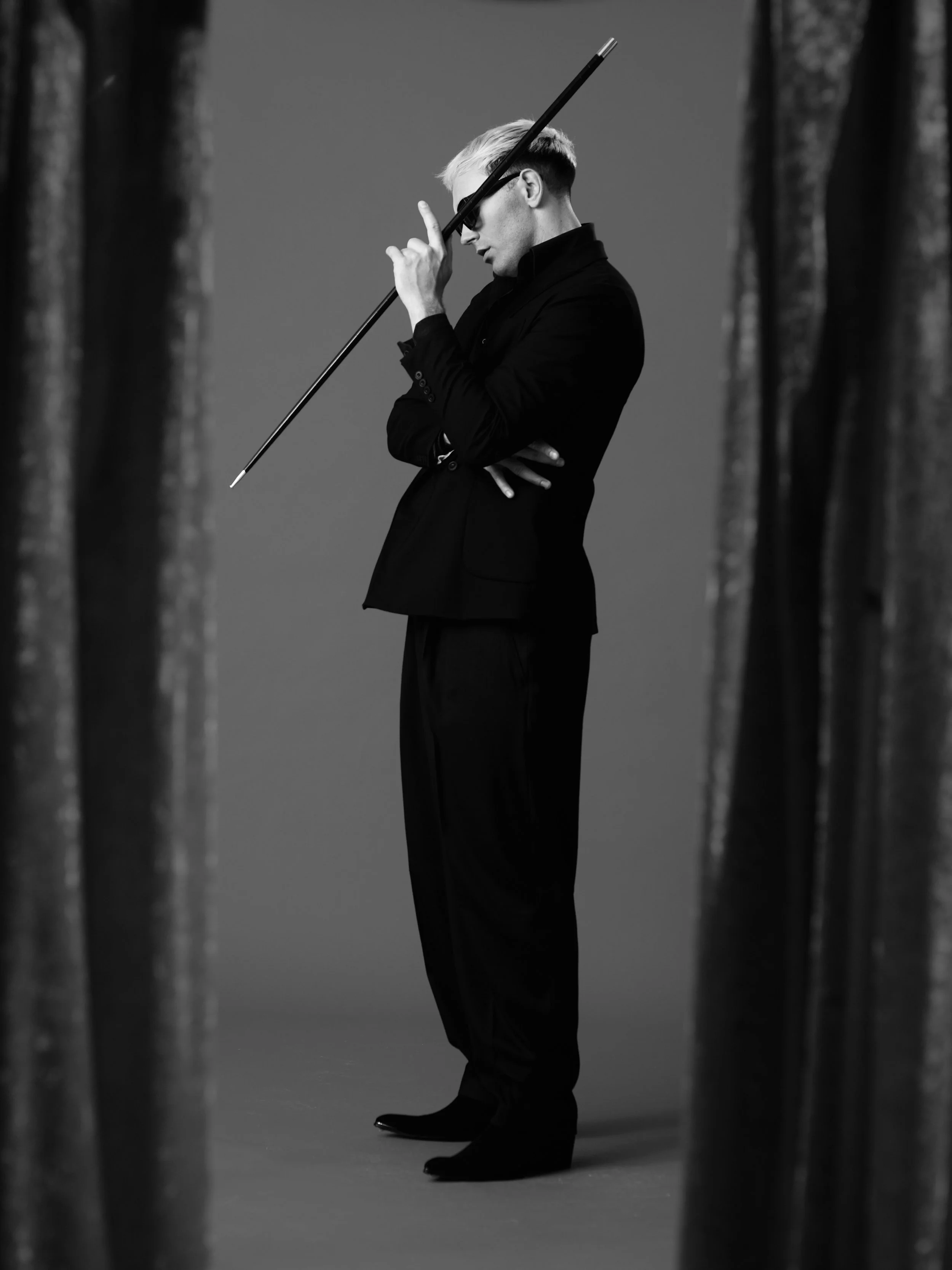 Black and white photo of a man with light-colored hair wearing a dark suit and sunglasses, holding a long object to his forehead, standing between two curtains or drapes.