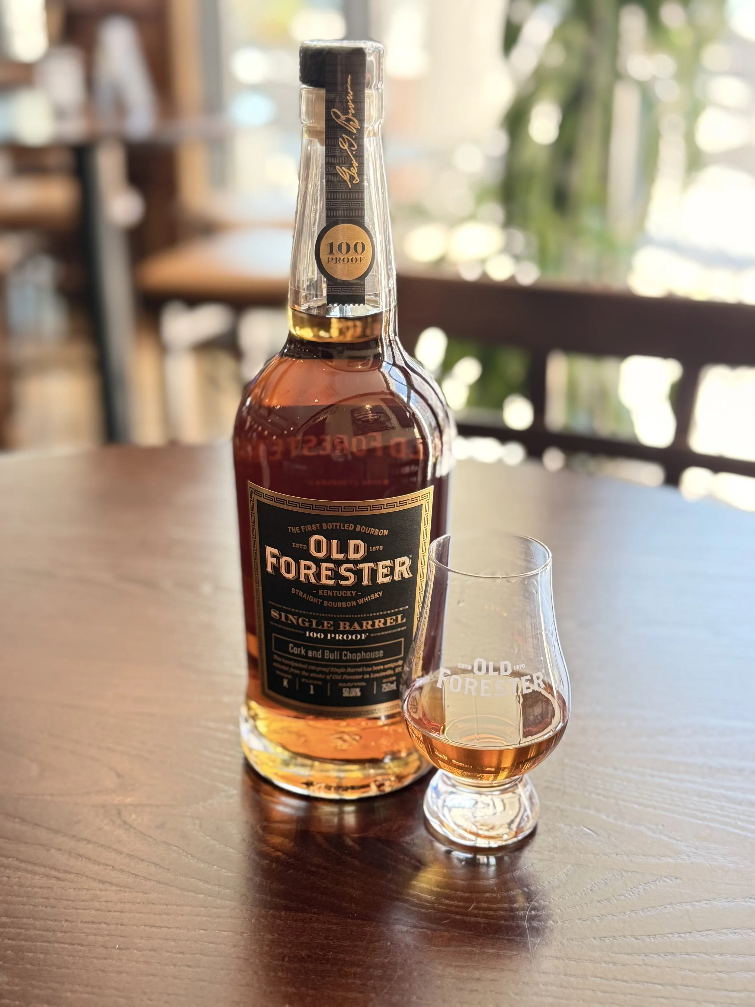 A bottle of Old Forester bourbon whiskey with a glass of bourbon on a wooden table.