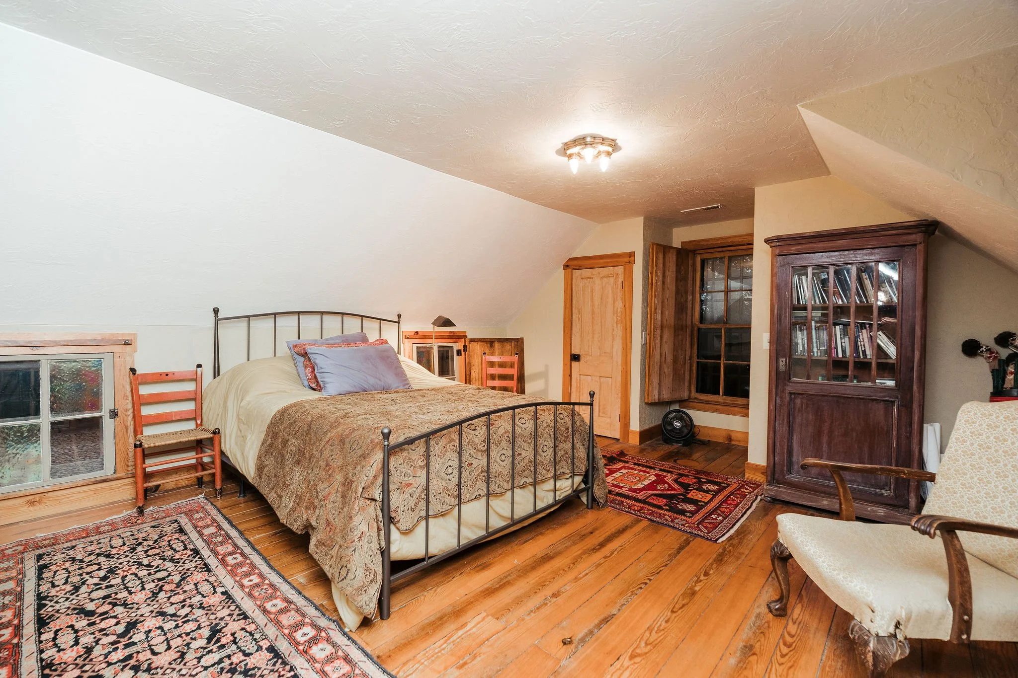 Bedroom with sloped ceilings, a metal bed with pillows and a quilt, wooden furniture including a wardrobe and nightstands, a patterned area rug, hardwood floor, and a window with wooden framing.