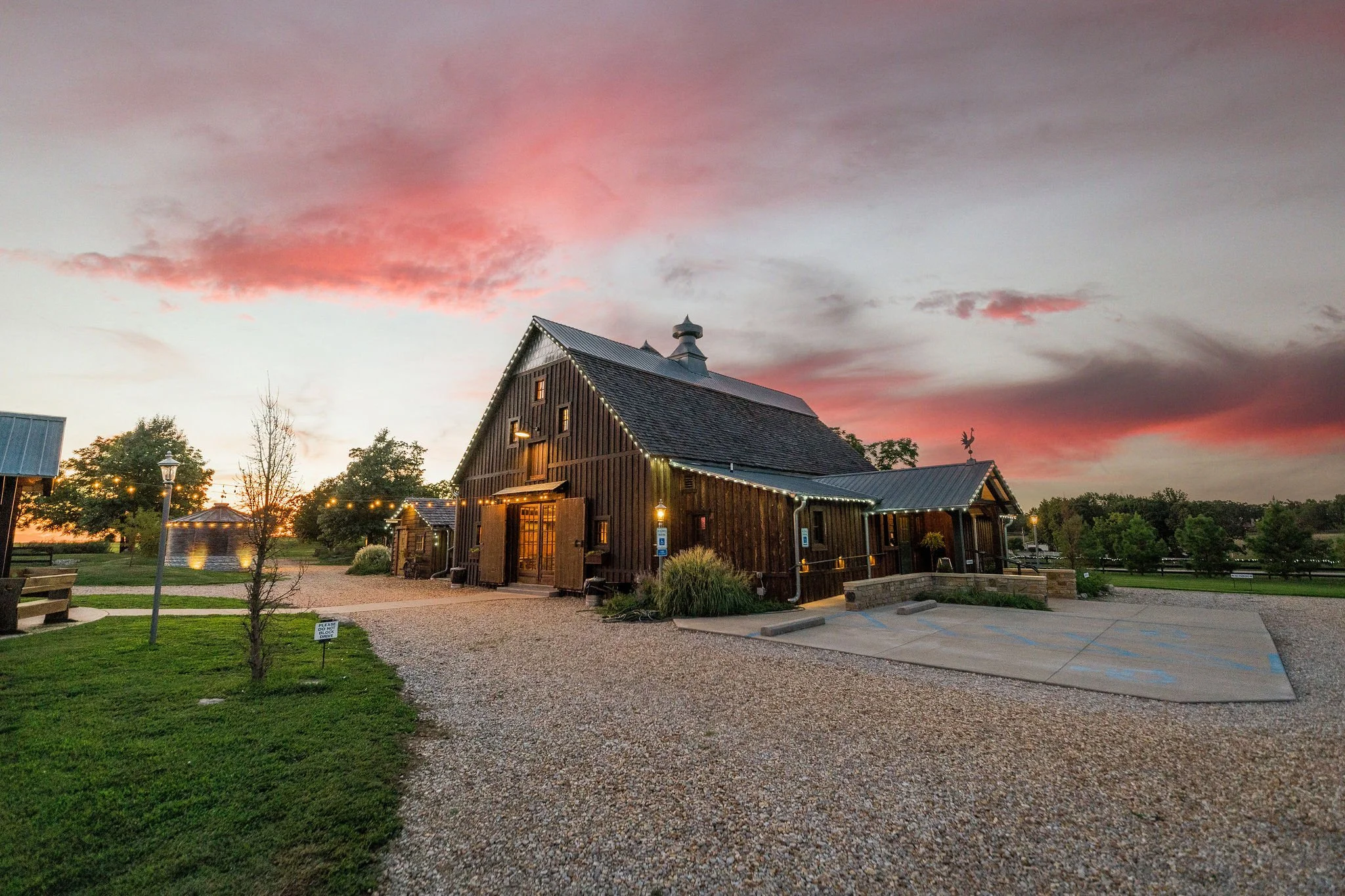 A rustic wooden barn with string lights at sunset, surrounded by a gravel driveway, green grass, trees, and a parking space for handicapped.