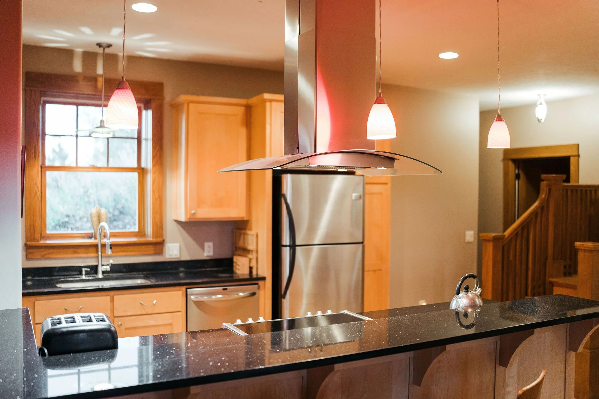 Kitchen with black granite countertops, stainless steel refrigerator, wooden cabinetry, double sink, hanging pendant lights, and a window over the sink