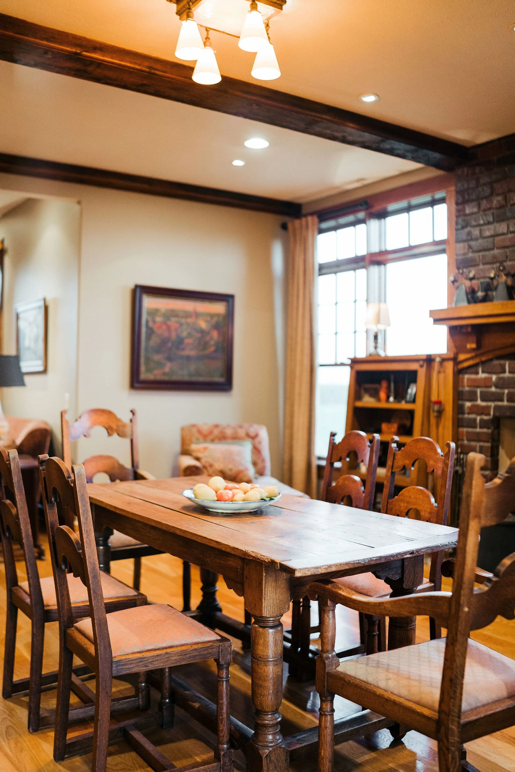 A rustic dining room with a wooden table and chairs, a brick fireplace, framed artwork, window with curtains, and ceiling lights.