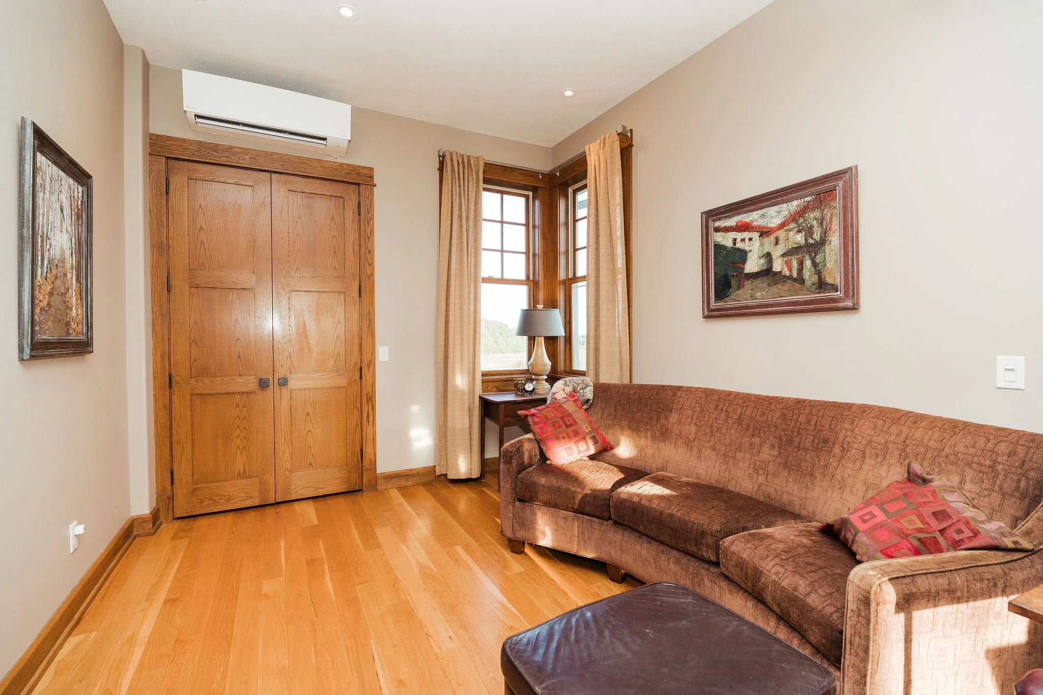 Living room with wooden flooring, a brown couch with patterned pillows, a wooden side table with a lamp, and framed artwork on the beige walls. A window with beige curtains and an air conditioning unit above a double wooden door are also visible.