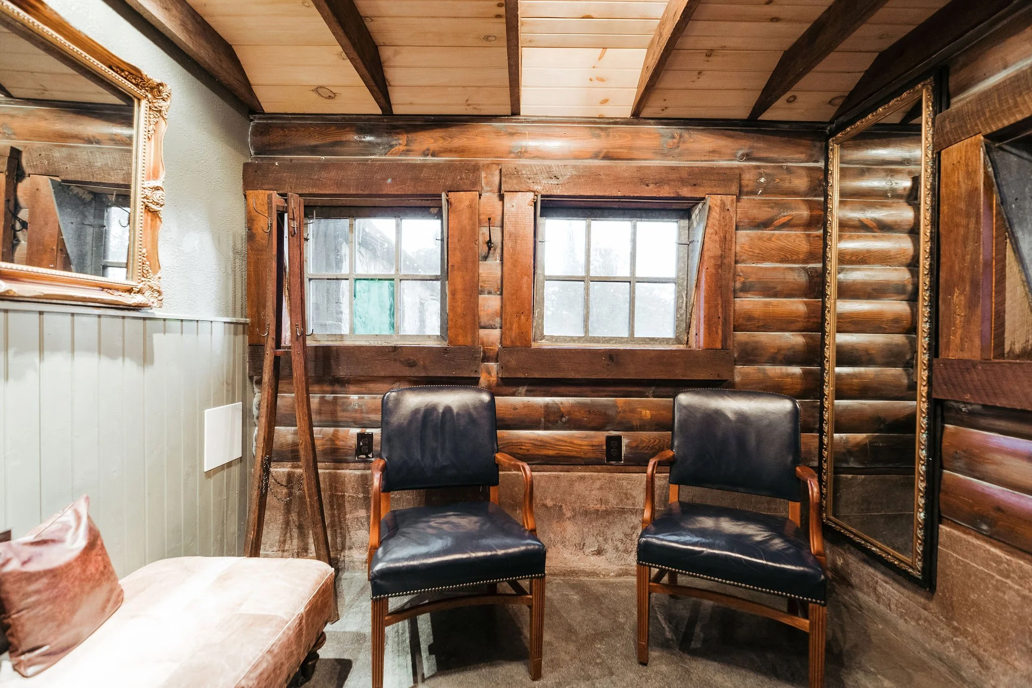 Room with wooden log walls, two small windows, two black leather chairs, a mirror, a beige upholstered chair with a pink cushion, and a wooden floor.