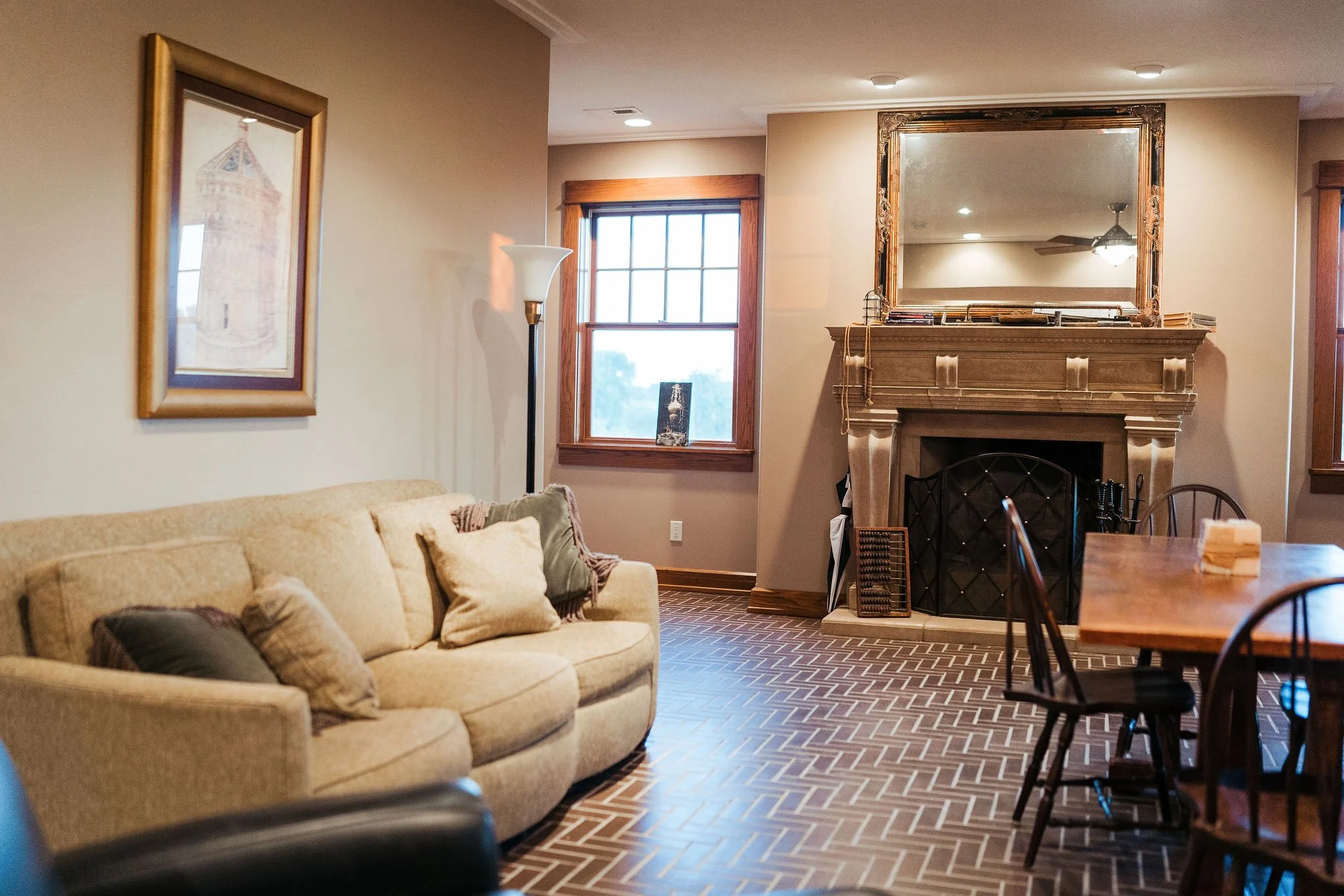 Living room with beige sofa, wooden dining table and chairs, fireplace with large gold-framed mirror, window with wood trim, and hardwood herringbone-patterned flooring.