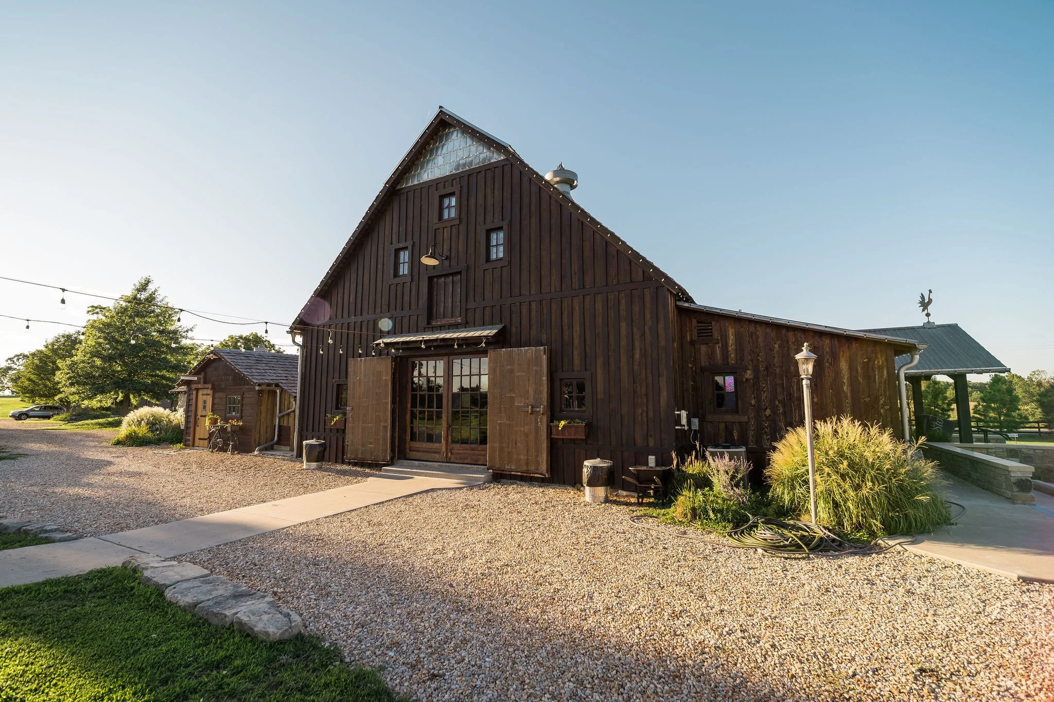 A dark wooden barn with double glass doors, set on a gravel area, with a pathway leading up to it. There are trees and a small grassy area nearby, with string lights hanging overhead, and outdoor decorative lights and plants around the barn in a rura