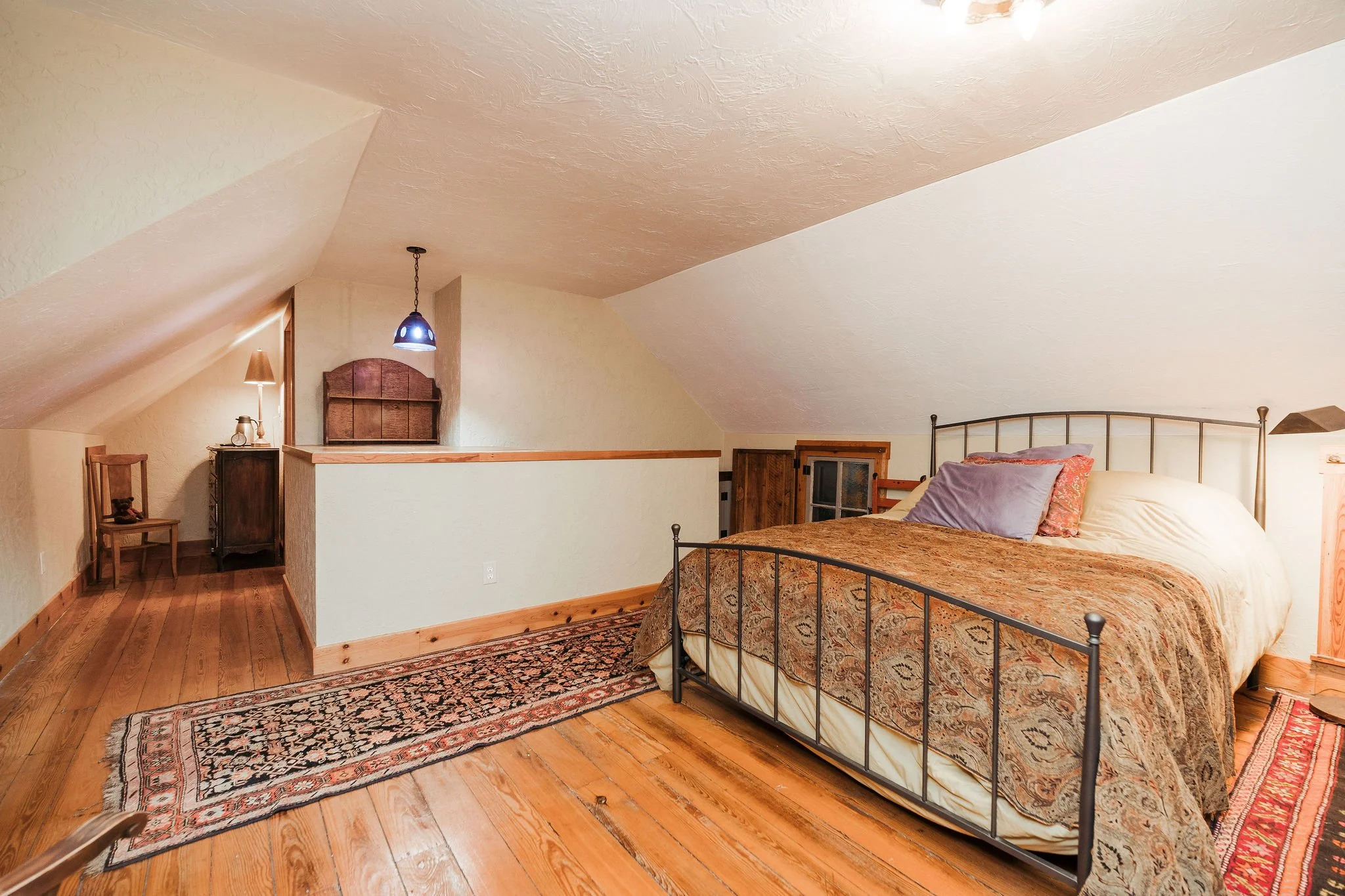 A cozy attic bedroom with a metal bed frame, patterned bedding, pillows, hardwood floors, a small window, and warm lighting.