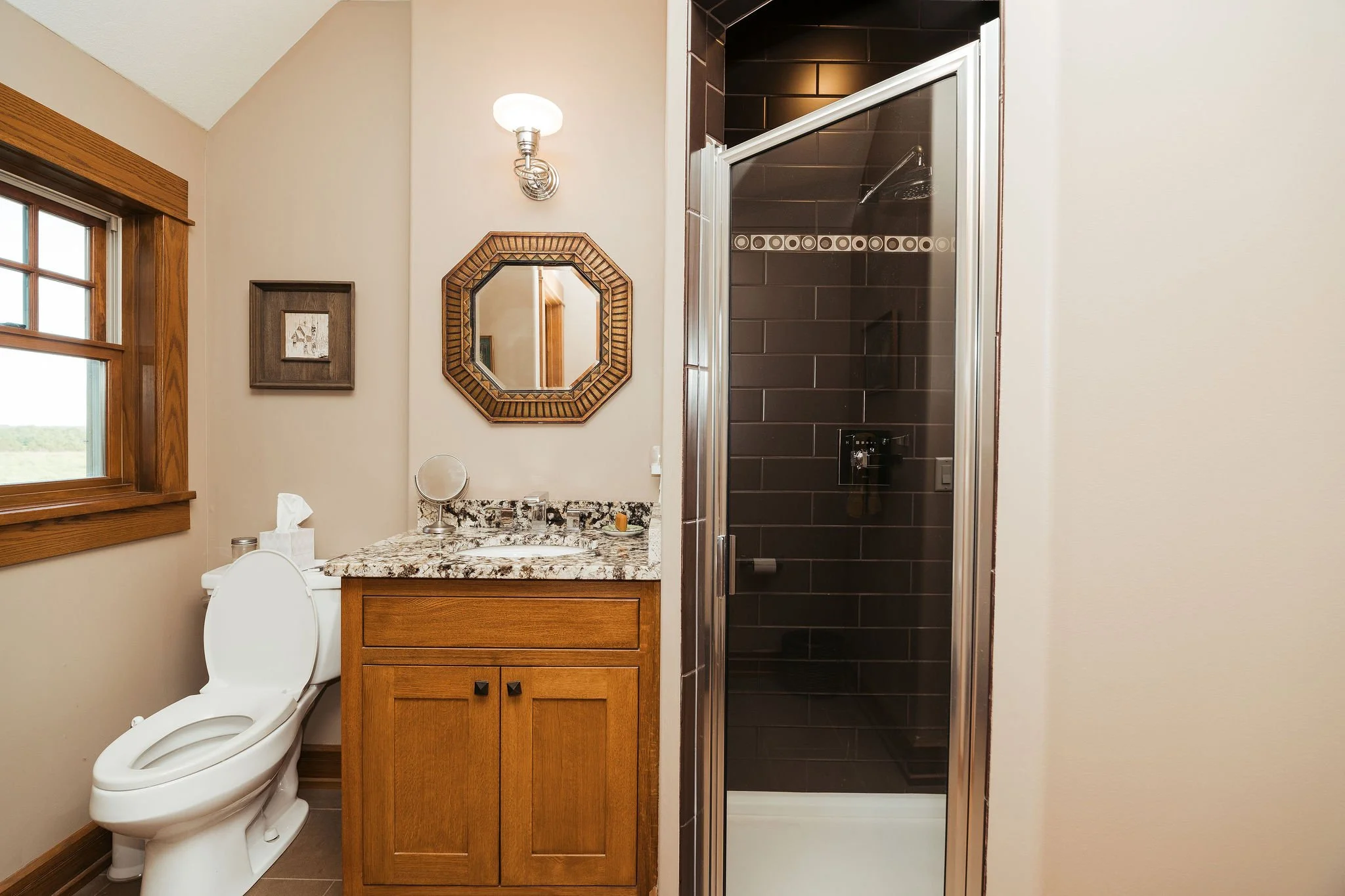 Bathroom with wooden window frame, a toilet, a wooden vanity with granite countertop, a small mirror, a wall light, and a shower with dark tiles and a glass door.