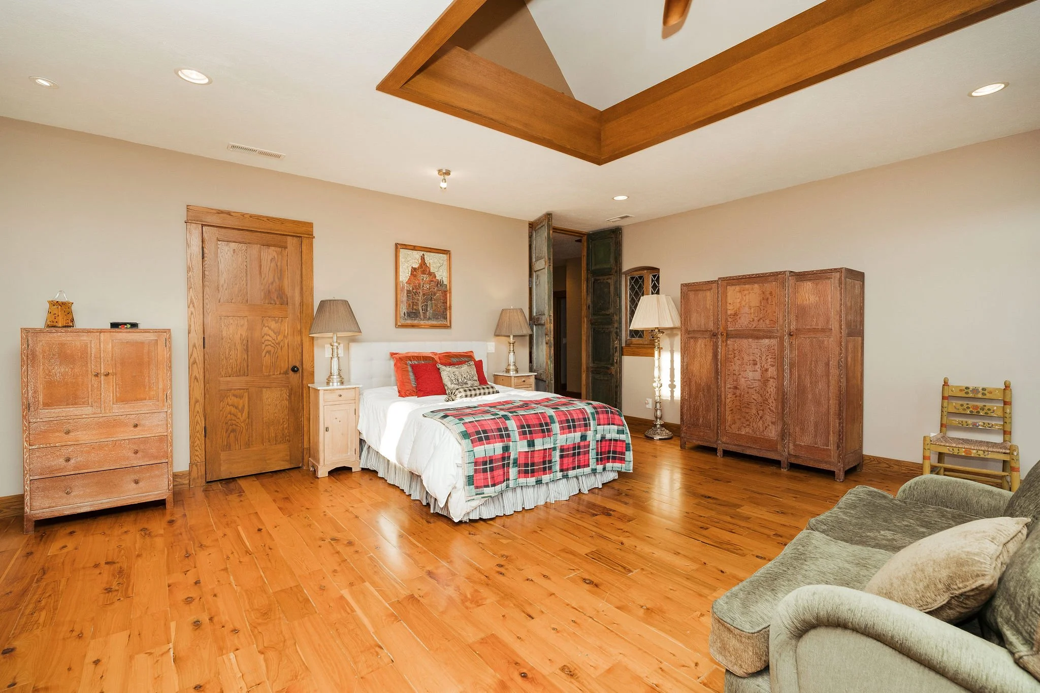 A bedroom with a wooden floor, a bed with a plaid blanket, and wooden furniture including a wardrobe, chest of drawers, and nightstand. There is a sofa to the right and a folding chair near the window.