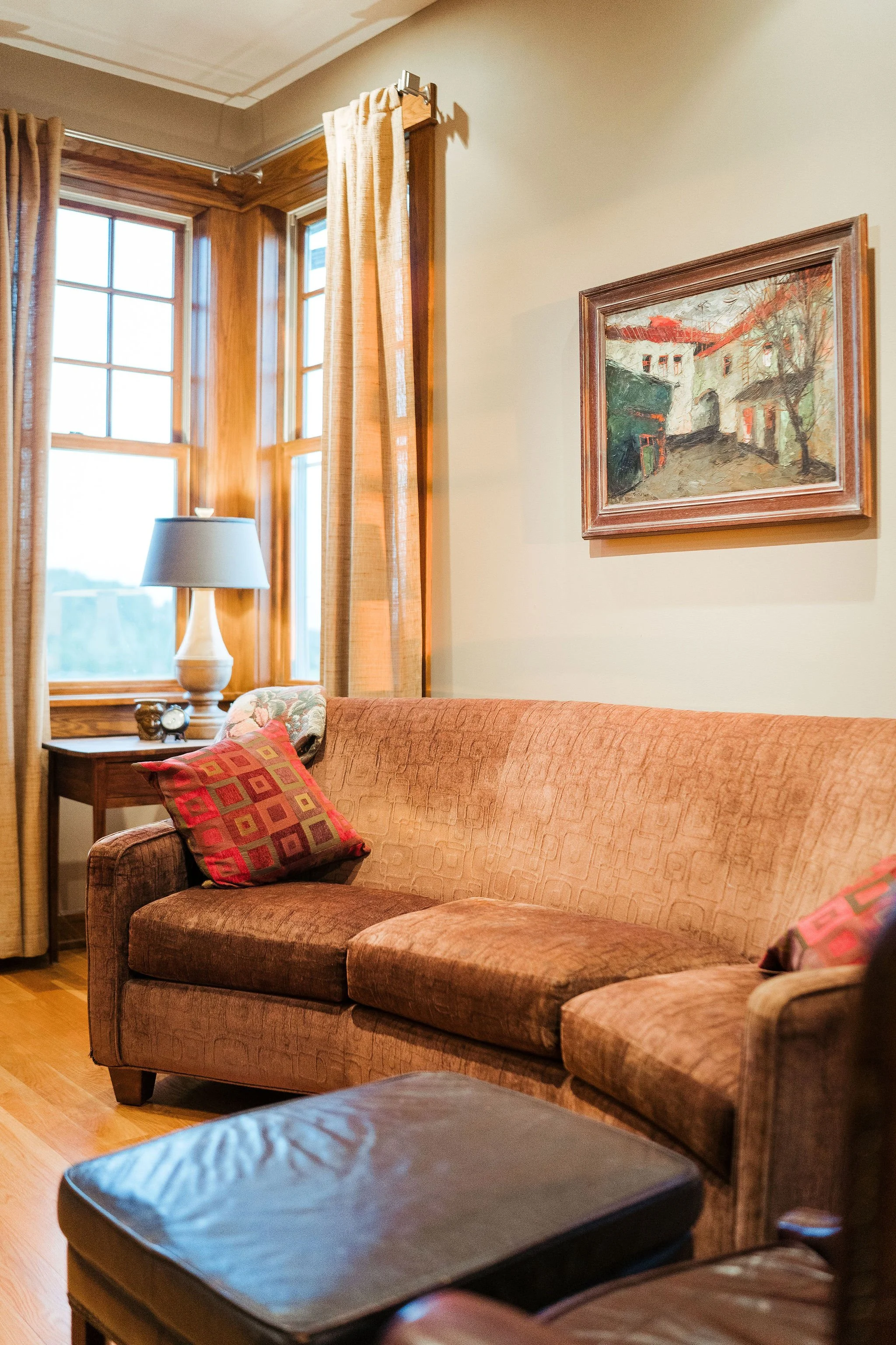 Cozy living room with a brown sofa, two decorative pillows, a wooden side table with a table lamp, a framed painting of a street scene, and windows with wooden trim and curtains.