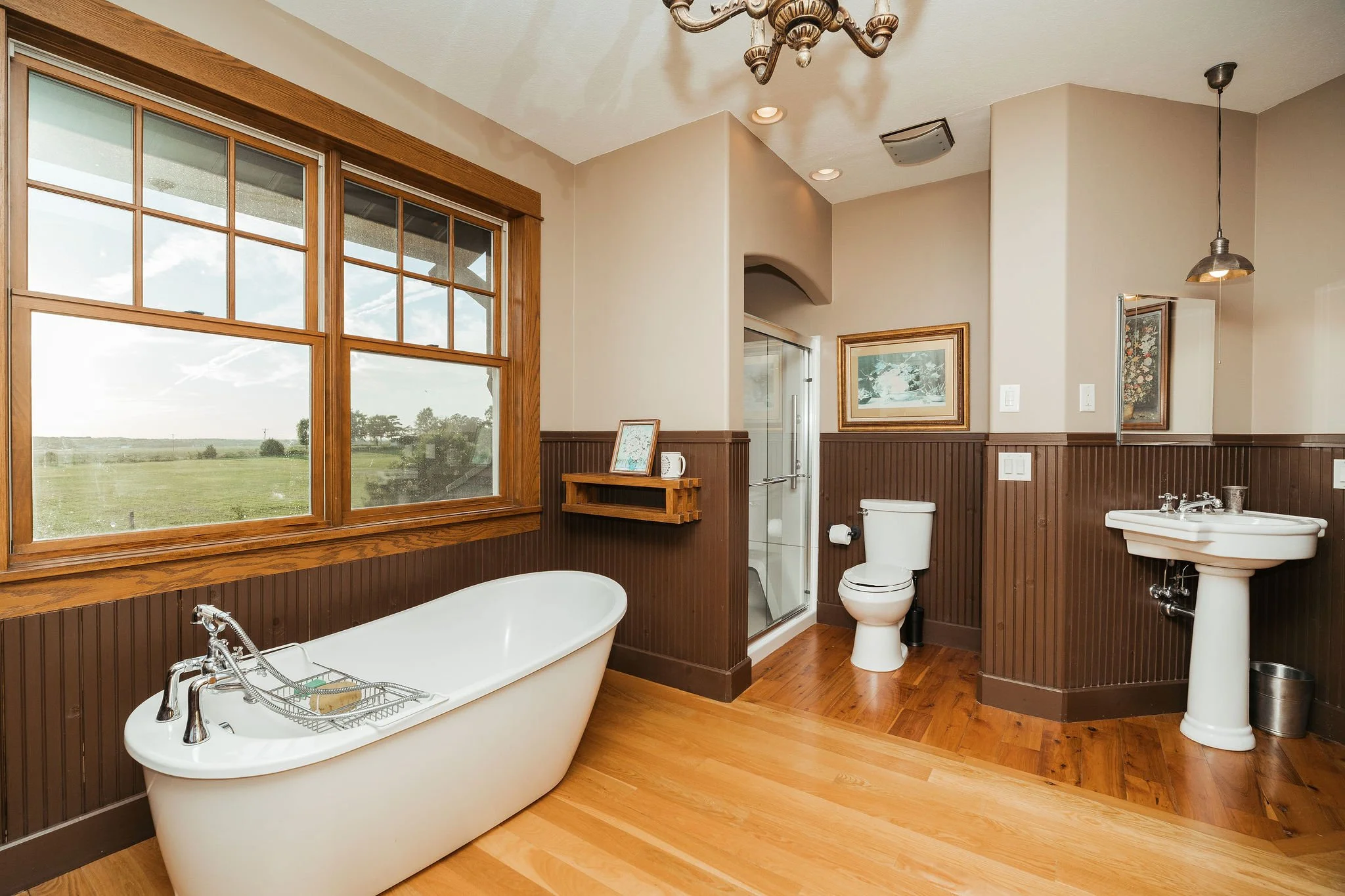 Bathroom with large window showing a green field, a white bathtub, a toilet, a pedestal sink, and framed artwork on beige and brown walls with wood paneling and hardwood floors.
