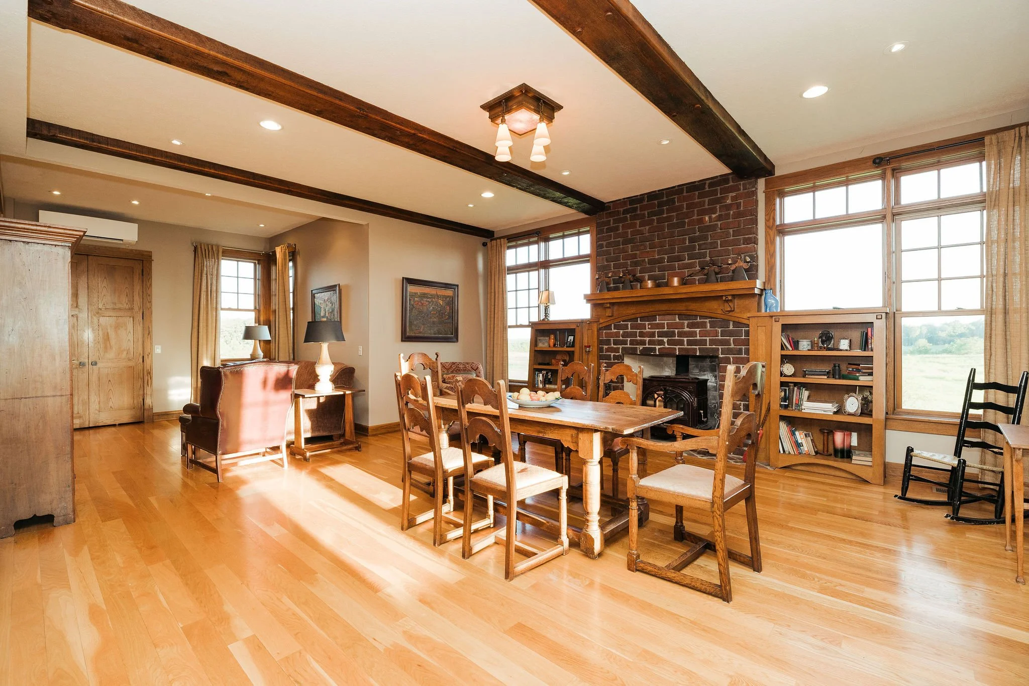 Bright living and dining area with wooden furniture, a brick fireplace, large windows, hardwood floors, and trim.