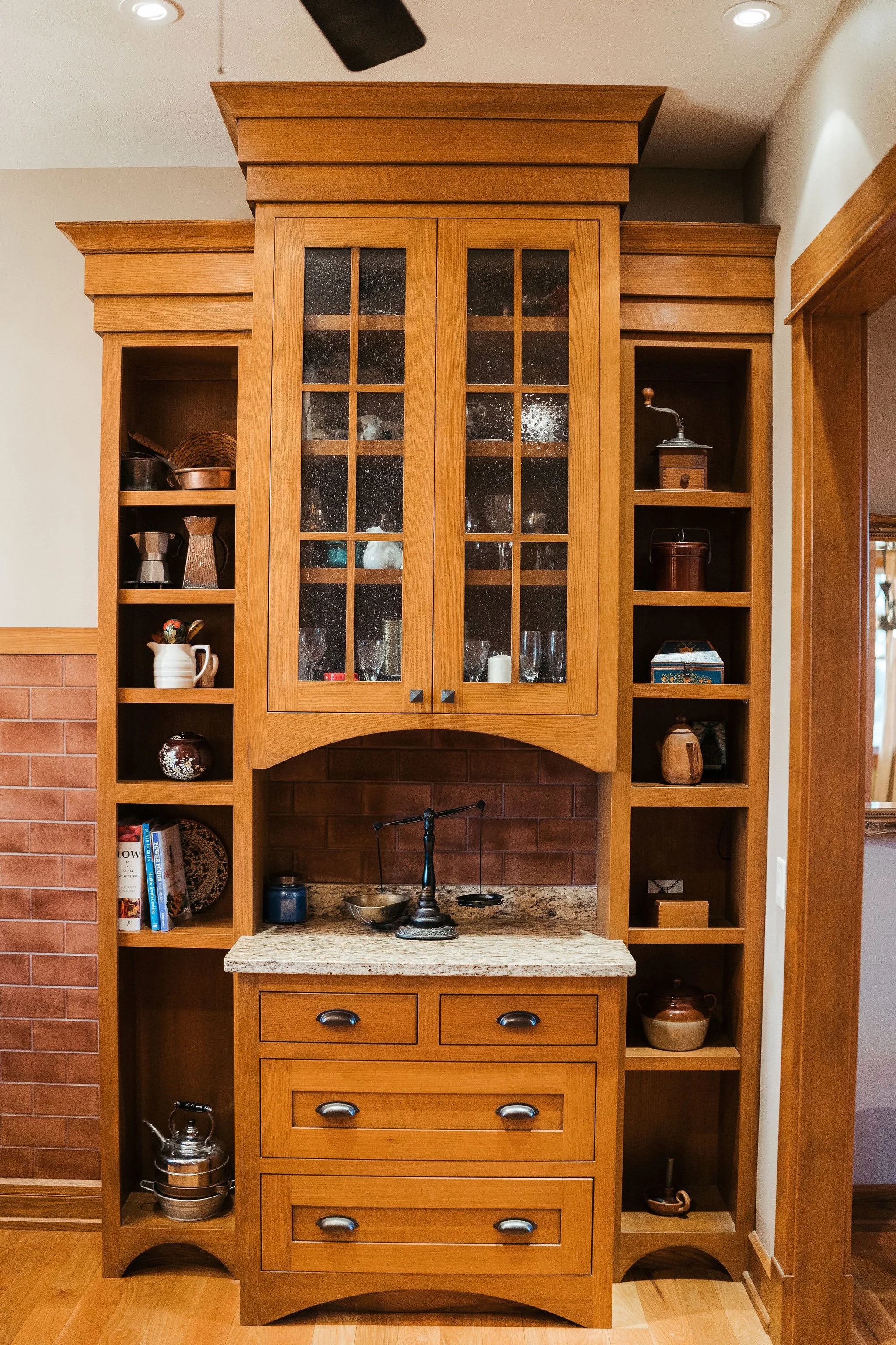 Wooden kitchen display cabinet with glass doors, containing glassware and decorative items, and a granite countertop with a vintage scale, set against a brick backsplash.
