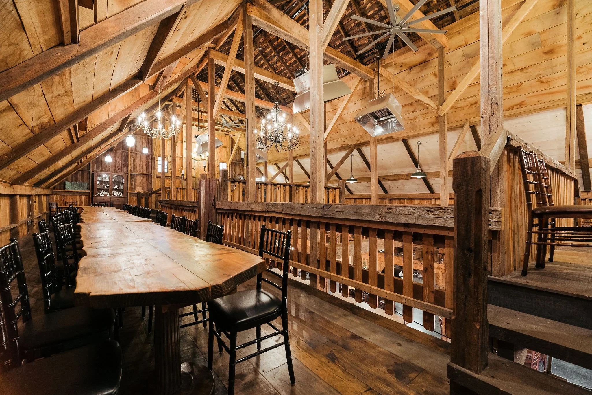 Interior of a rustic wooden barn with a long dining table, black chairs, chandeliers, and warm lighting.