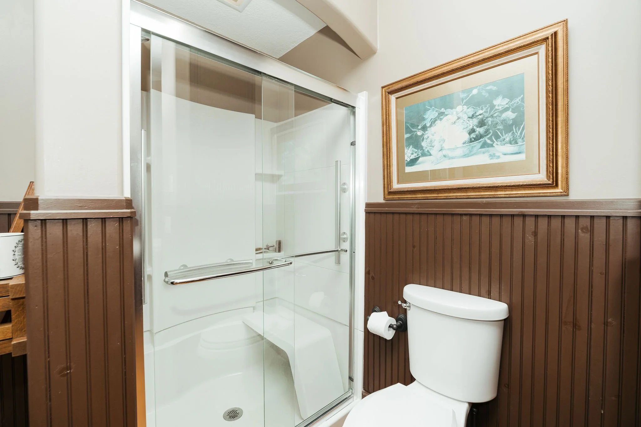 Bathroom with a glass shower stall, a white toilet, a framed painting of fruit on the wall, and brown wooden wall paneling.