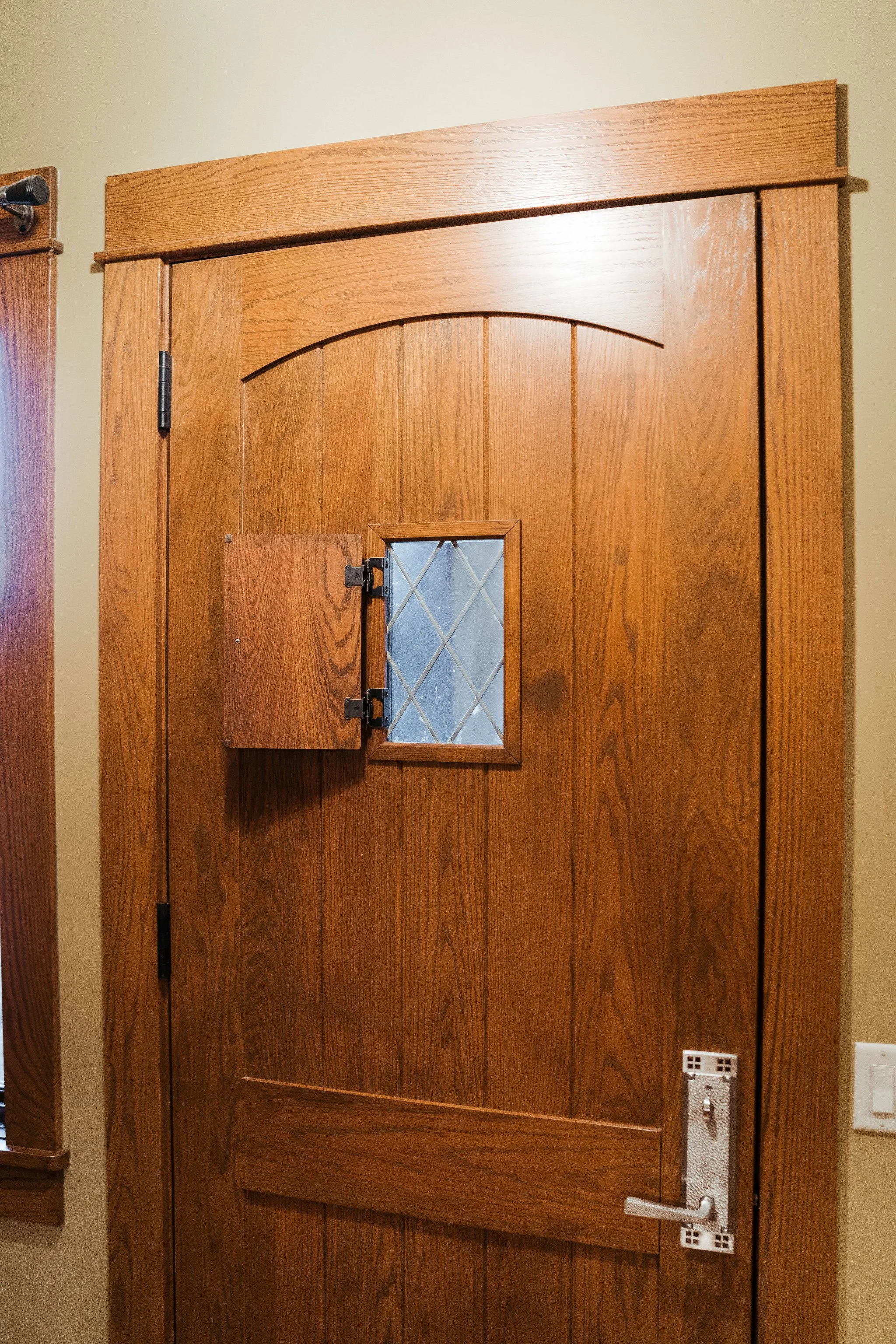 Wooden interior door with small diamond-patterned window and partially open small wooden hatch, set in a room with light-colored walls.