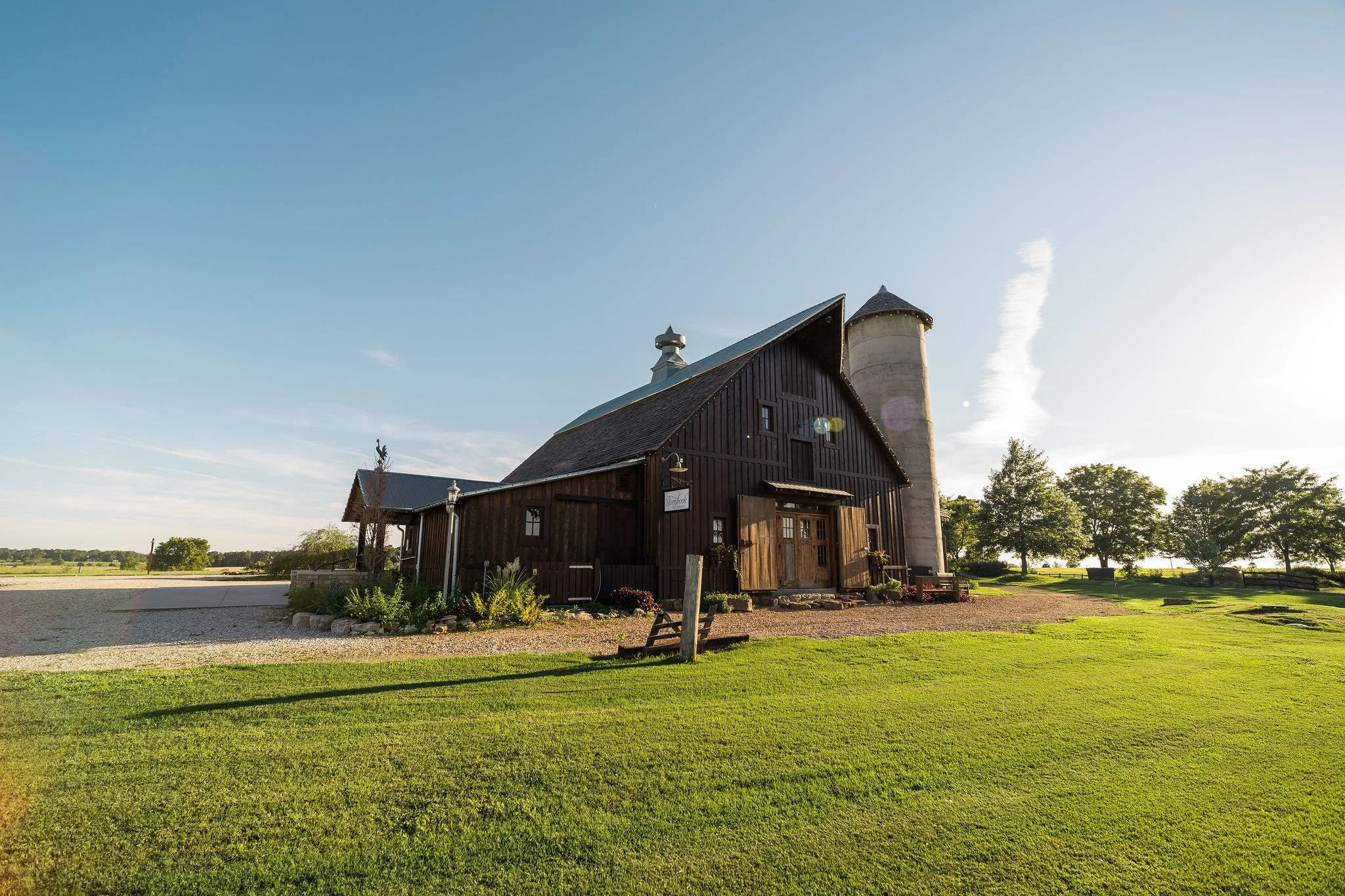 A large rustic barn with wooden siding and a metal roof, surrounded by lush green grass and trees, with a silo attached, against a bright blue sky.