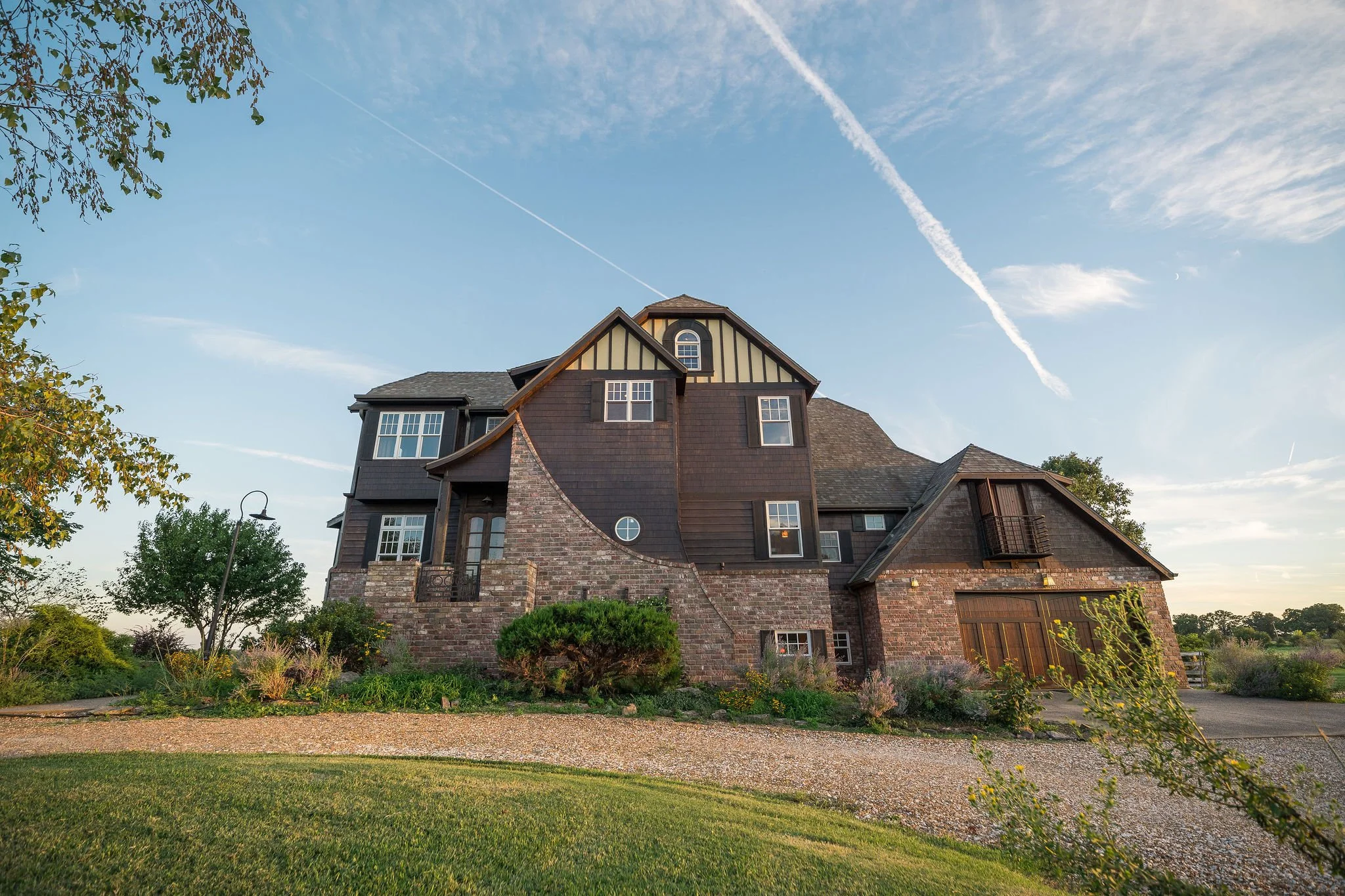 A large house with a mixture of brick and dark wood siding, multiple windows, and a sloped roof, surrounded by greenery and a gravel driveway under a blue sky with wispy clouds.