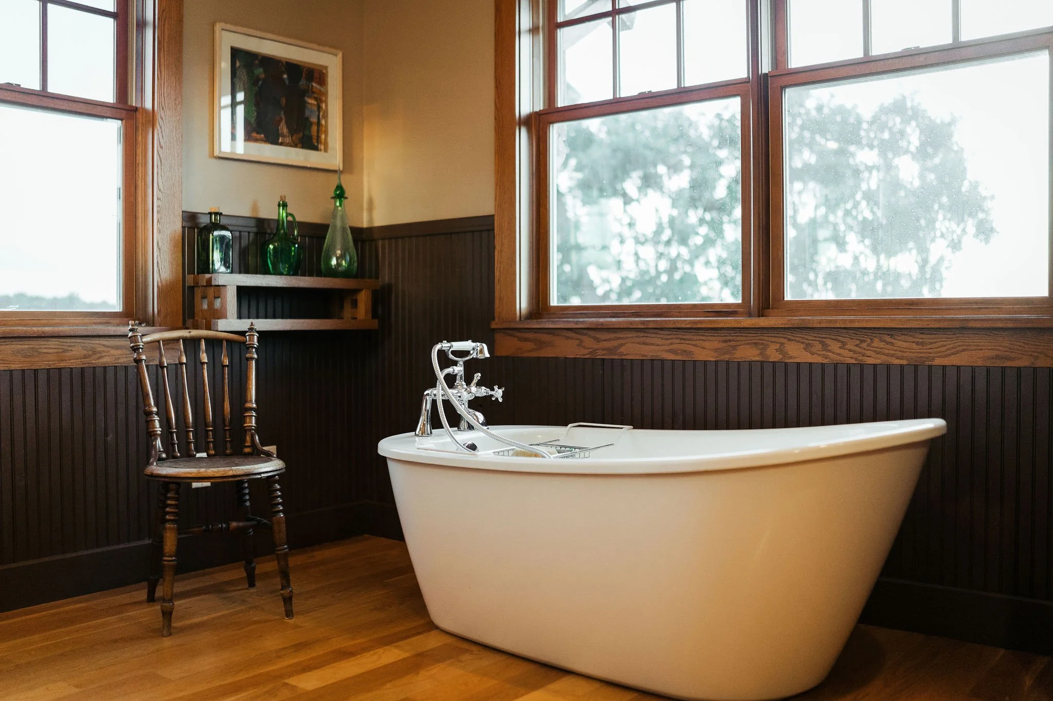 A vintage bathroom with a standalone bathtub, a wooden chair, and large windows allowing natural light, decorated with green glass bottles on a shelf and framed artwork on the wall.