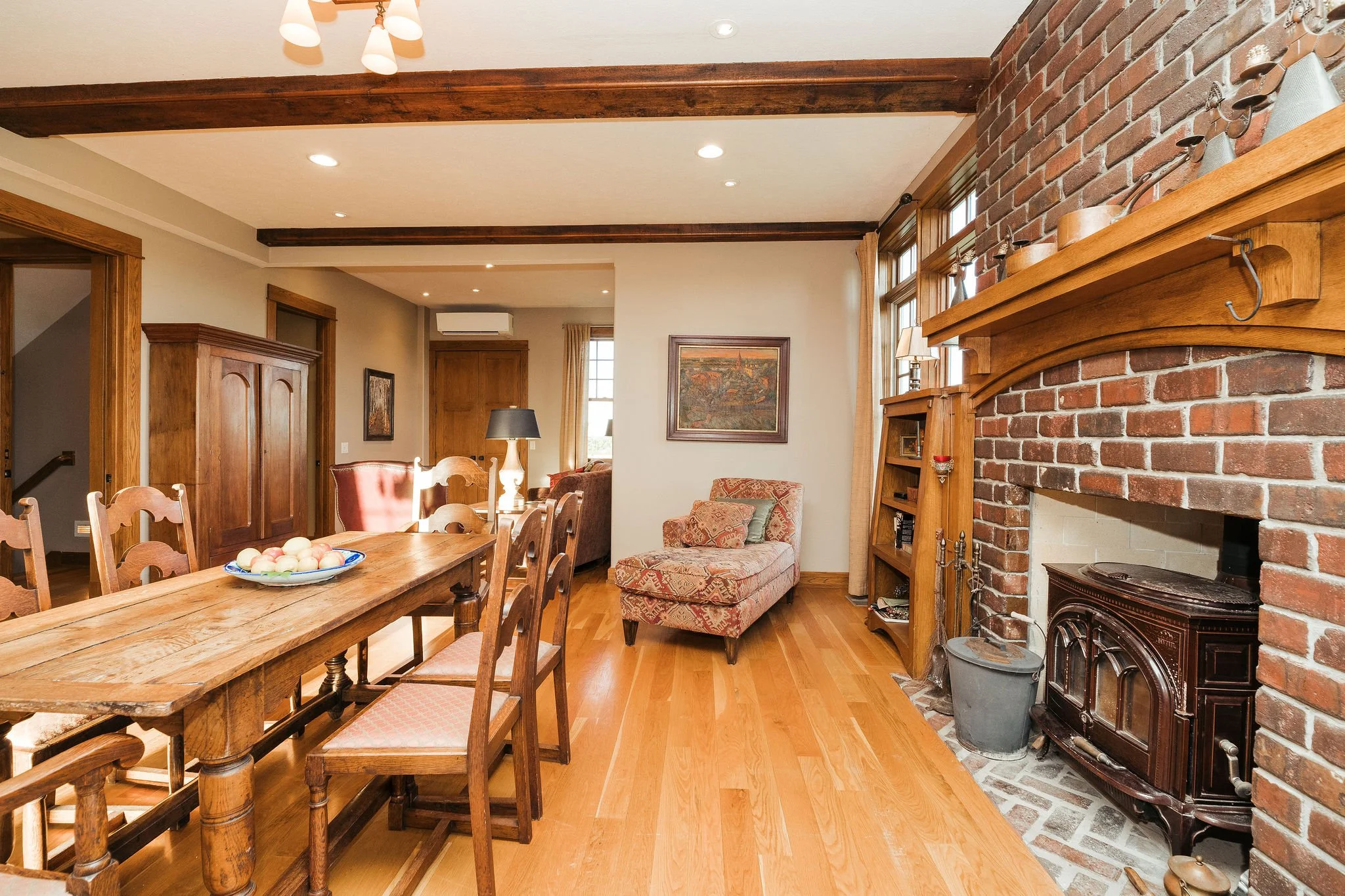 Living room featuring a wooden dining table with chairs, an armchair near the window, a brick fireplace with a wooden mantle, wooden bookshelves, hardwood floors, and large windows with curtains.