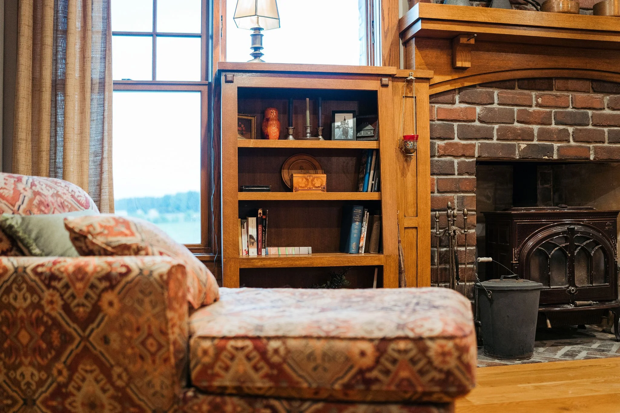 Living room corner featuring a floral upholstered sofa, a window with curtains, a wooden bookshelf with decorative items and books, a brick fireplace, and a black wood stove.