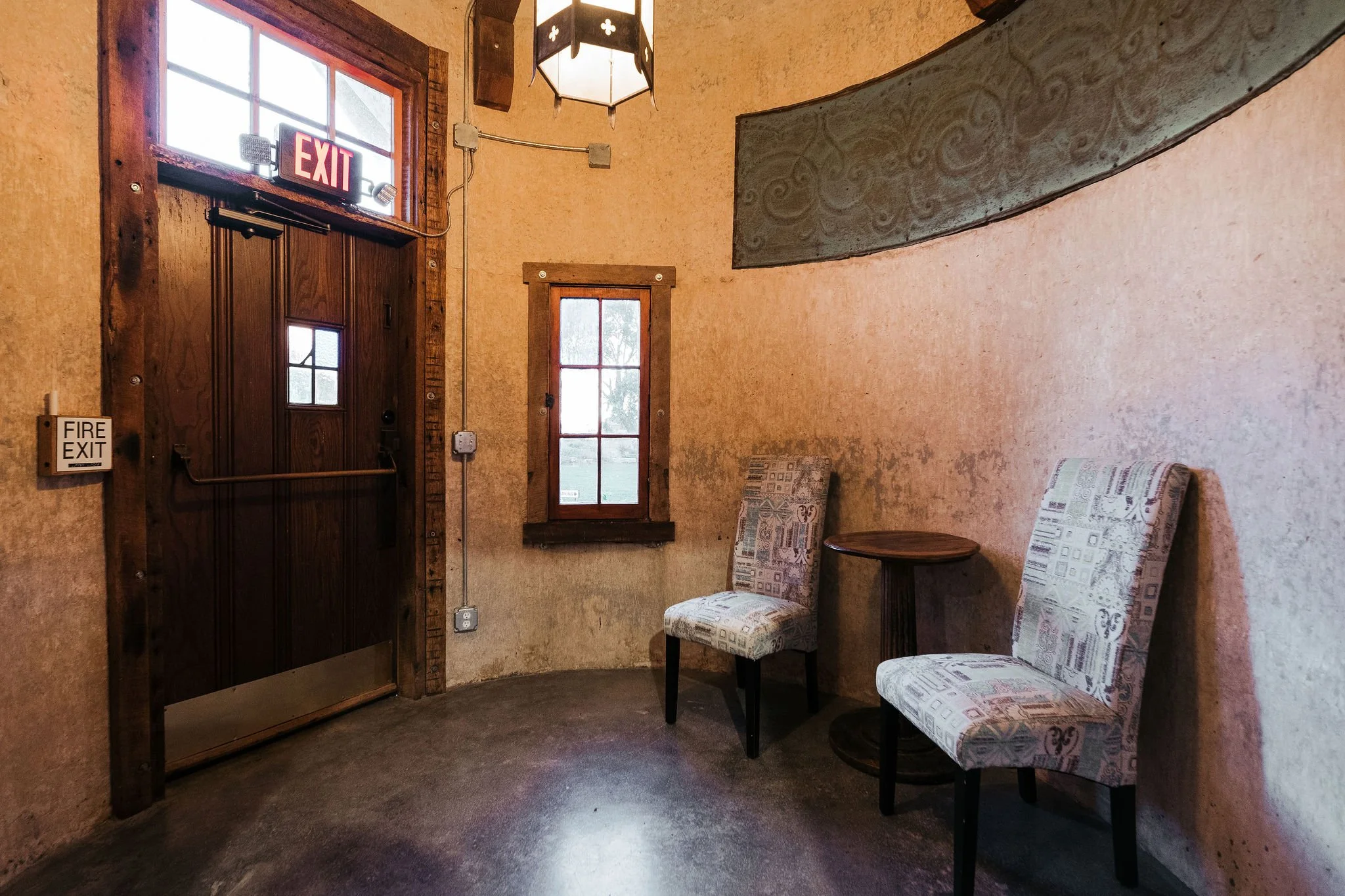Interior of a cozy room with two patterned chairs, a small round table, and windows with wooden frames, including an emergency exit door and fire exit sign.
