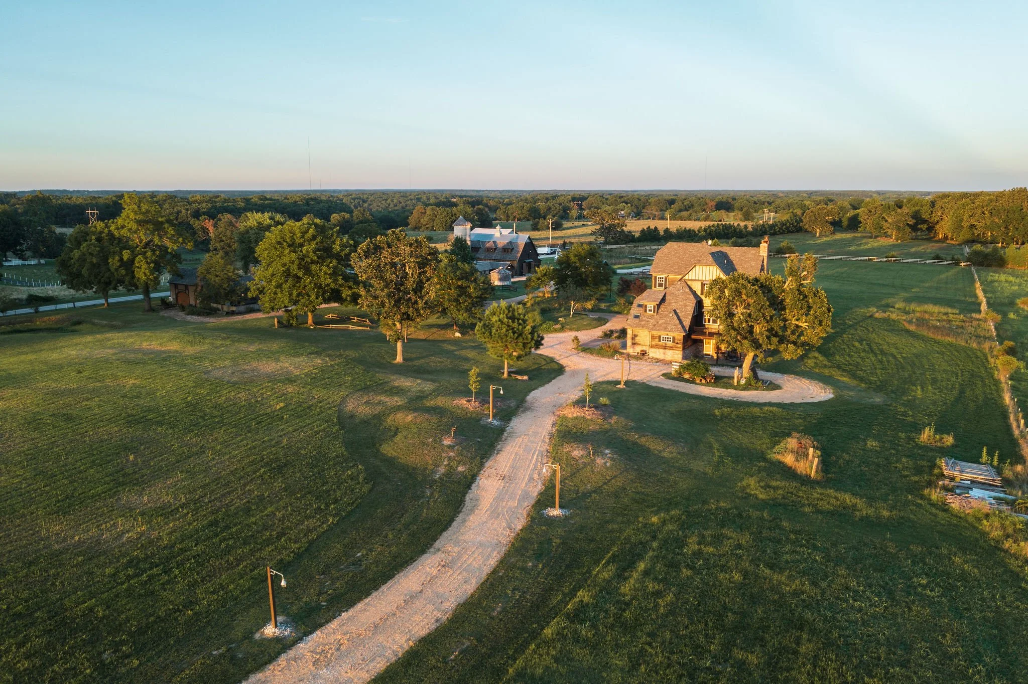 Aerial view of a rural property with a gravel driveway, multiple trees, green fields, and several buildings under a clear sky.