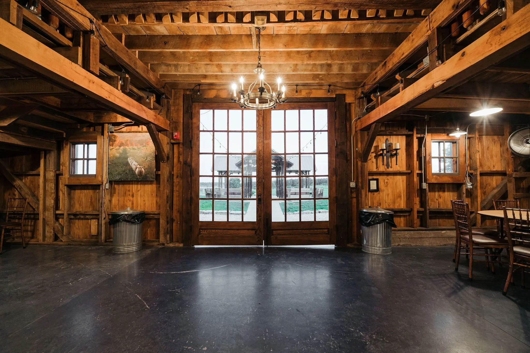 Interior of a rustic wooden barn with large glass doors showing a barn and fields outside, a chandelier hanging from the ceiling, and wooden chairs and tables along the walls.