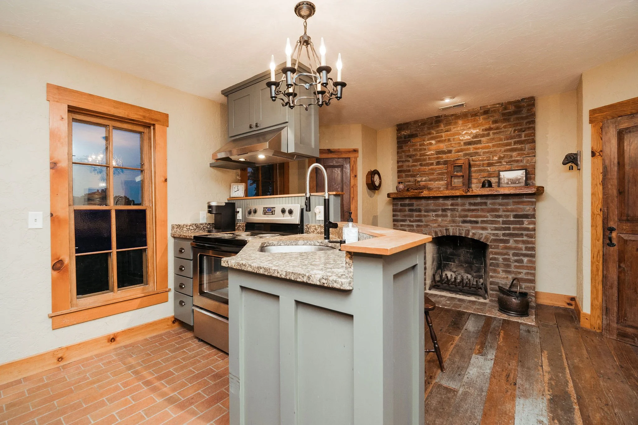 Kitchen with brick fireplace, wooden floor, gray cabinets, granite countertops, and a window with wooden trim.