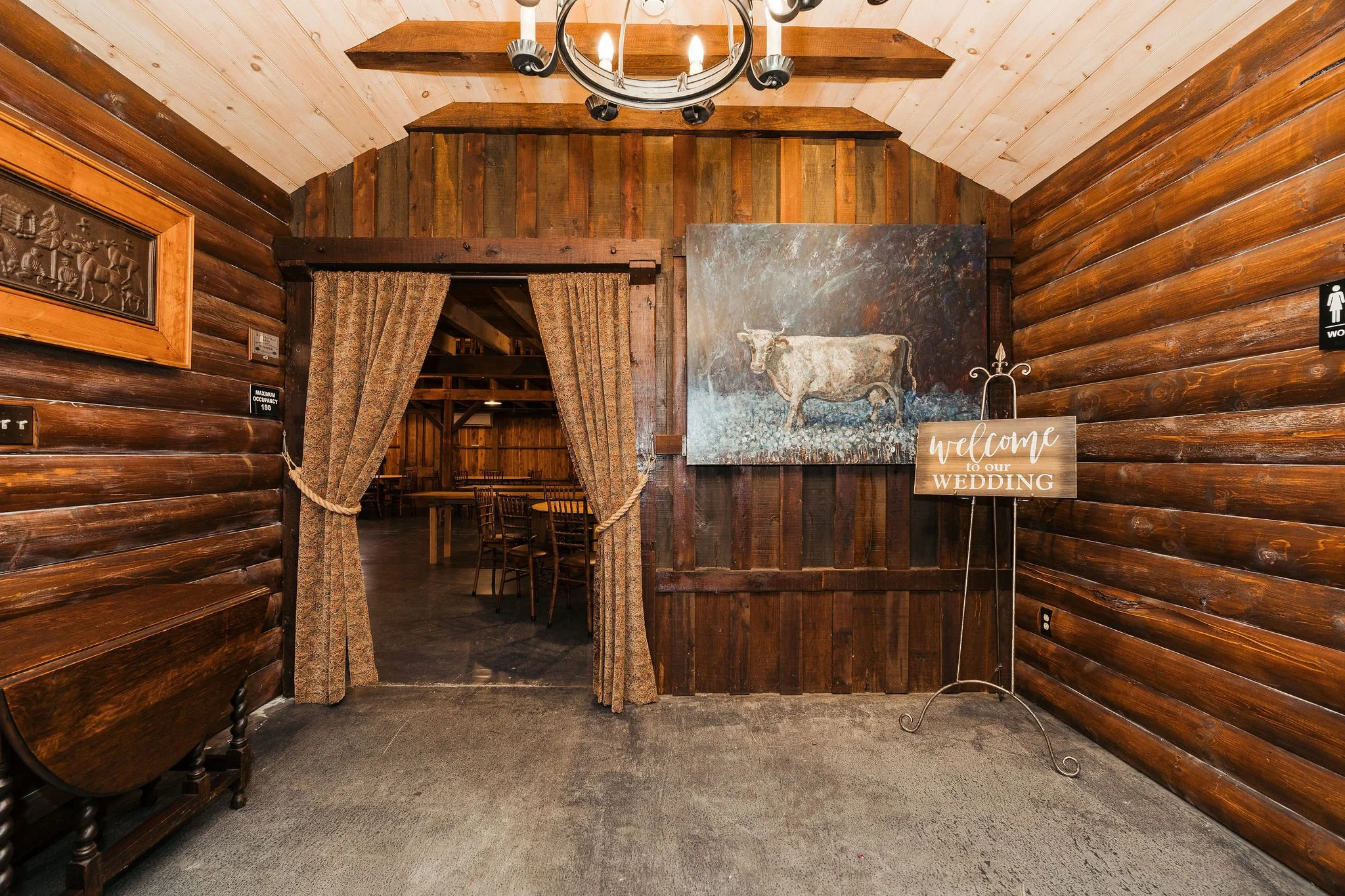 Wooden interior with a doorway covered by curtains, a large painting of a cow on the wall, and a sign welcoming guests to a wedding, indicating the setting is a rustic wedding venue.