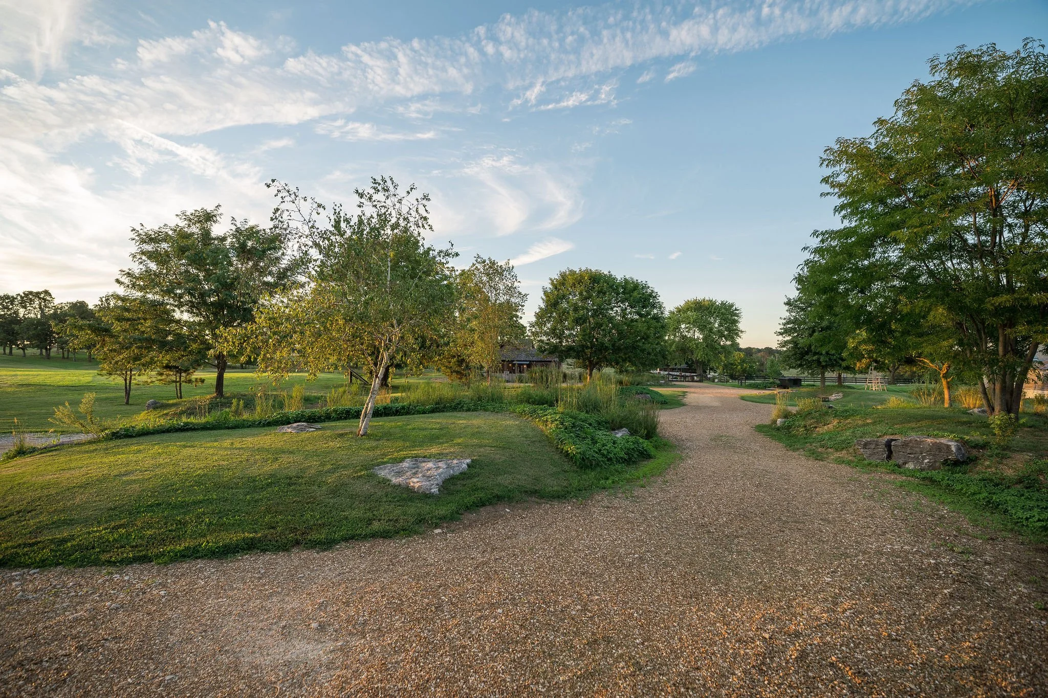 A peaceful parkscene with a gravel pathway winding through green grass and trees under a partly cloudy sky.