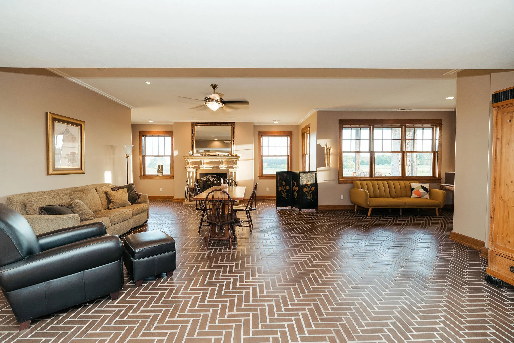 Spacious living room with beige walls, hardwood trim, brown herringbone patterned tile flooring, large windows, sofa with pillows, black leather armchair with footrest, dining table with chairs, TV on a stand, fireplace with mirror, decorative screen