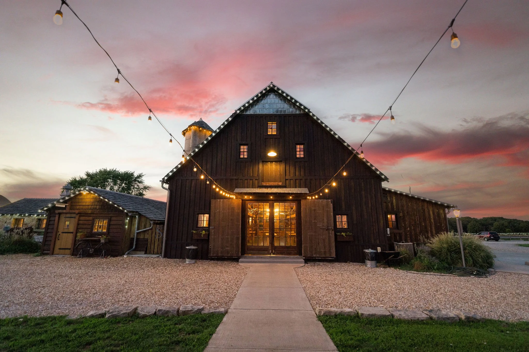 A rustic barn with string lights hanging across the front, during sunset with pink and purple clouds in the sky. Gravel path leads to open double doors.