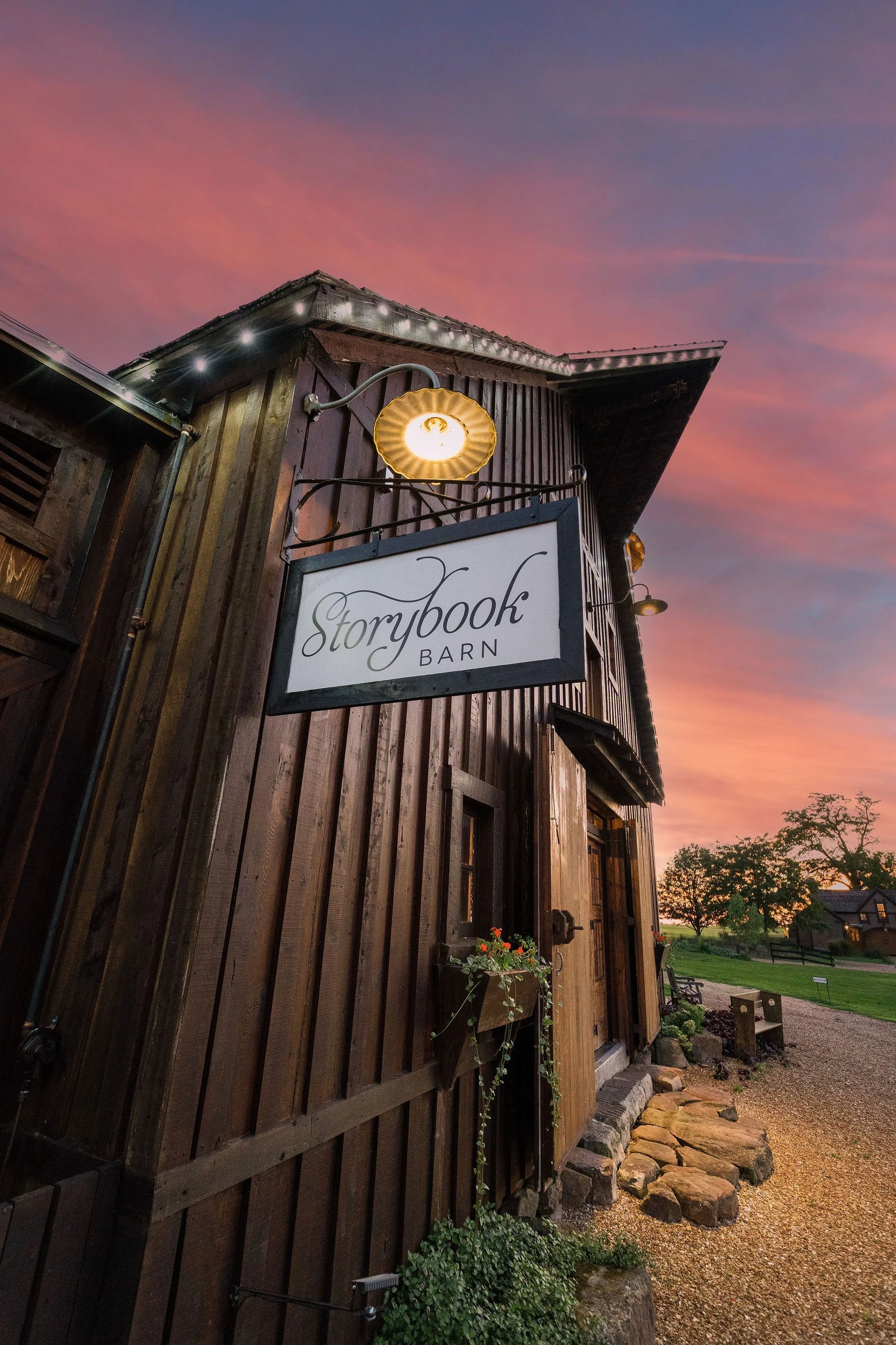 A rustic wooden barn with a sign that reads 'Storybook Barn' hanging outside, illuminated by a warm light at sunset.