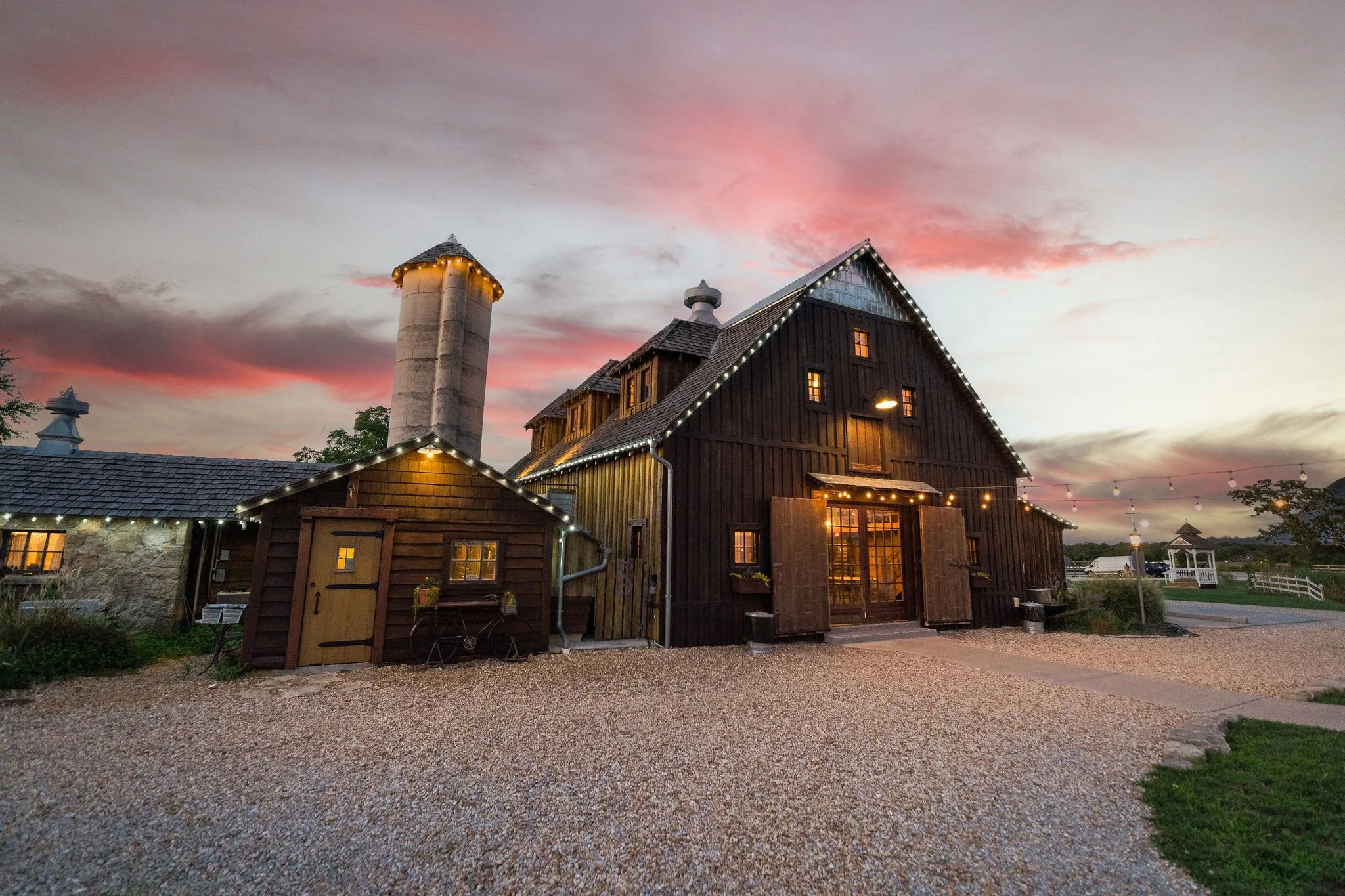 A rustic barn with string lights and a silo at sunset, with a gravel driveway and a gazebo in the background.