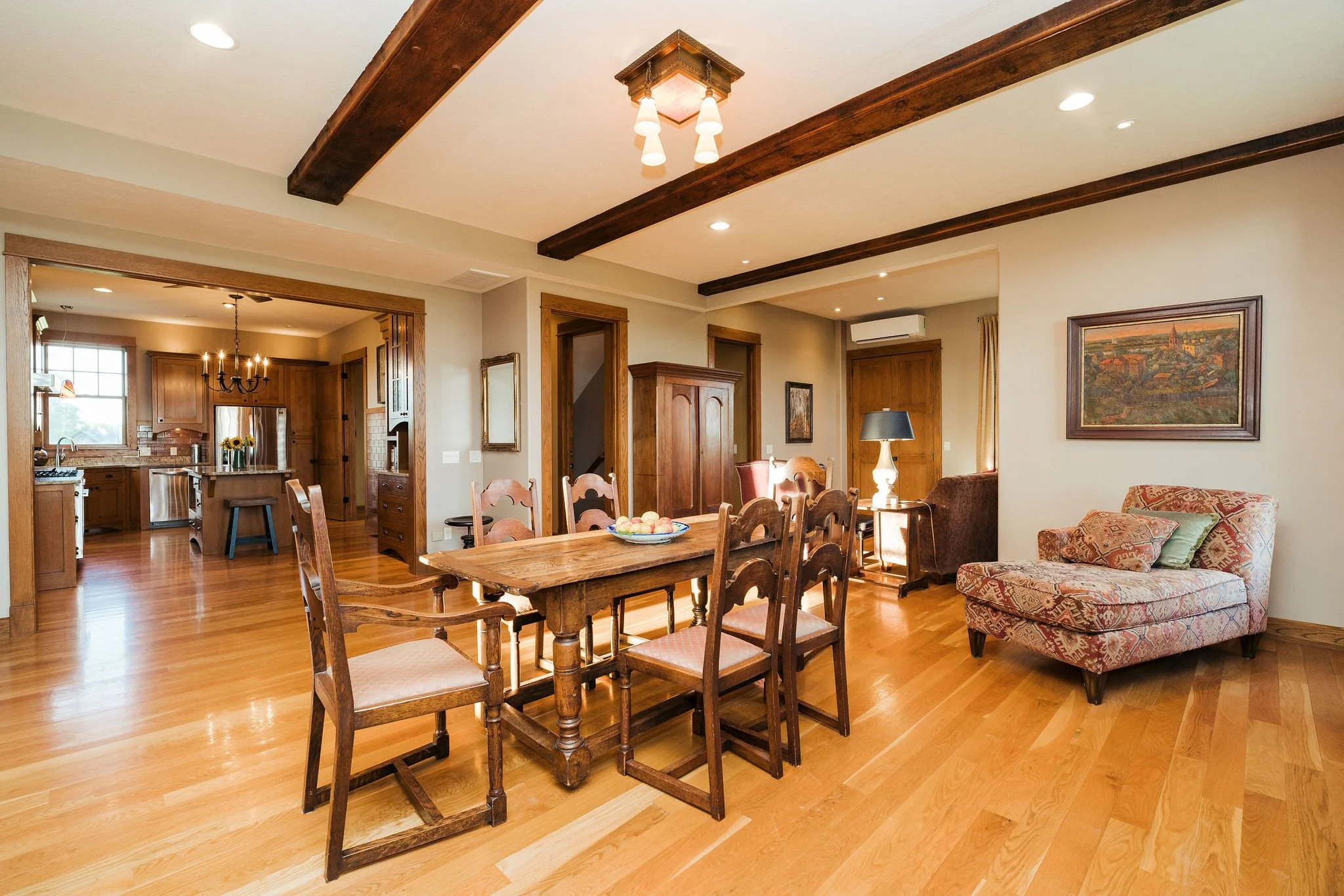 Open-concept living and dining area with hardwood floors, wooden beams on the ceiling, and a kitchen in the background. The dining table has eight chairs, and there is a cushioned armchair with pillows and a side table with a black lamp nearby. Artwo