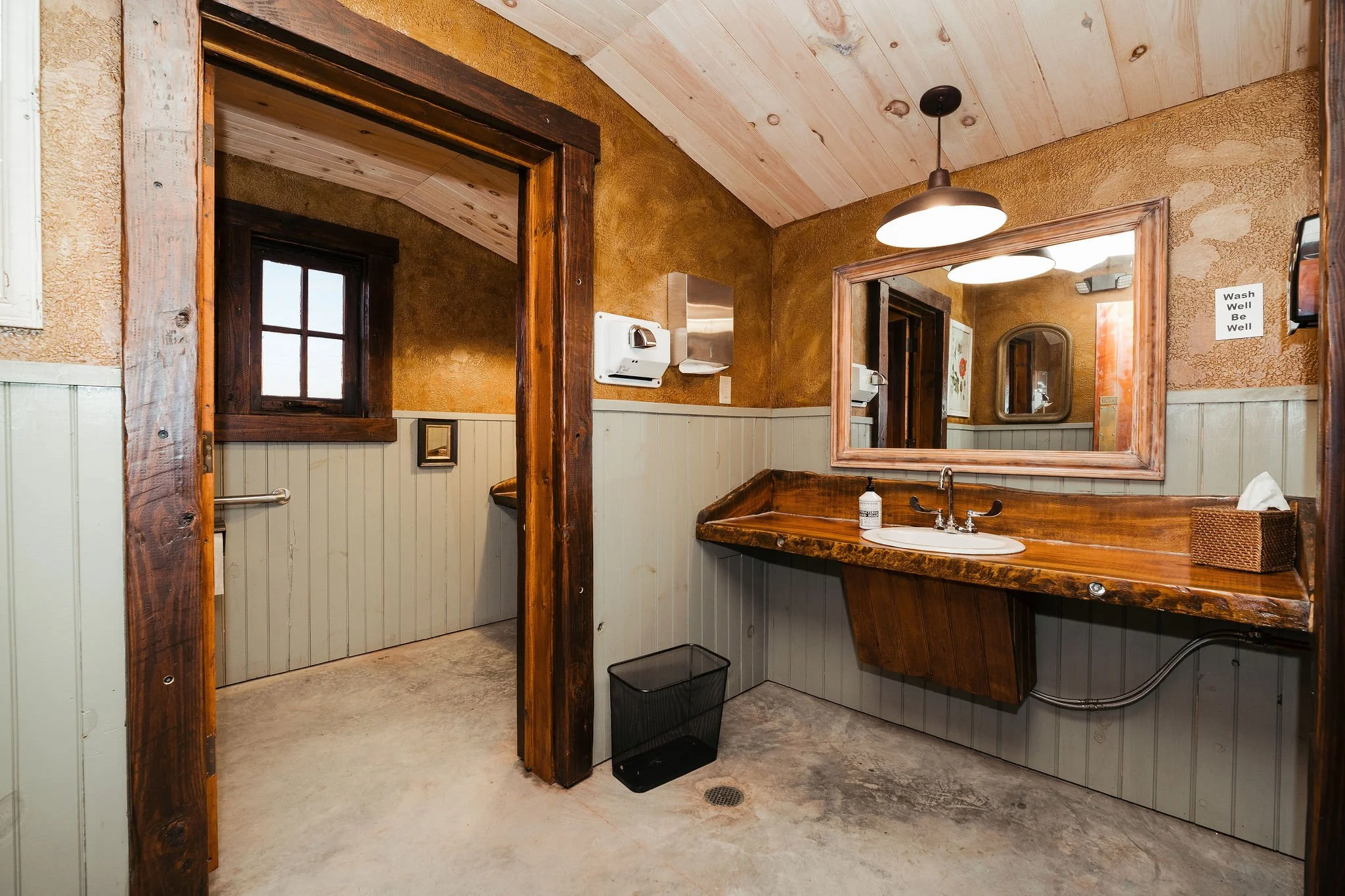 A rustic bathroom with beige beadboard wainscoting, a wooden vanity with a large mirror, a black trash bin, a paper towel dispenser, a soap dispenser, and a sign that says 'Wash Well Be Well'. An open doorway reveals a small room with a window and a