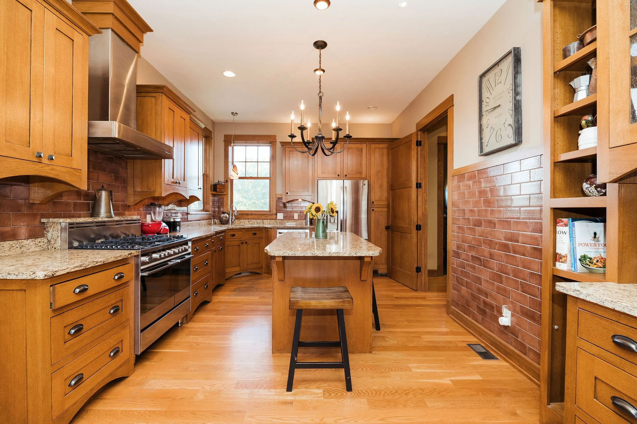 Wooden kitchen with granite countertops and brick backsplash, central island with a sunflower vase, chandelier overhead, stainless steel appliances, and open shelving with cookbooks.