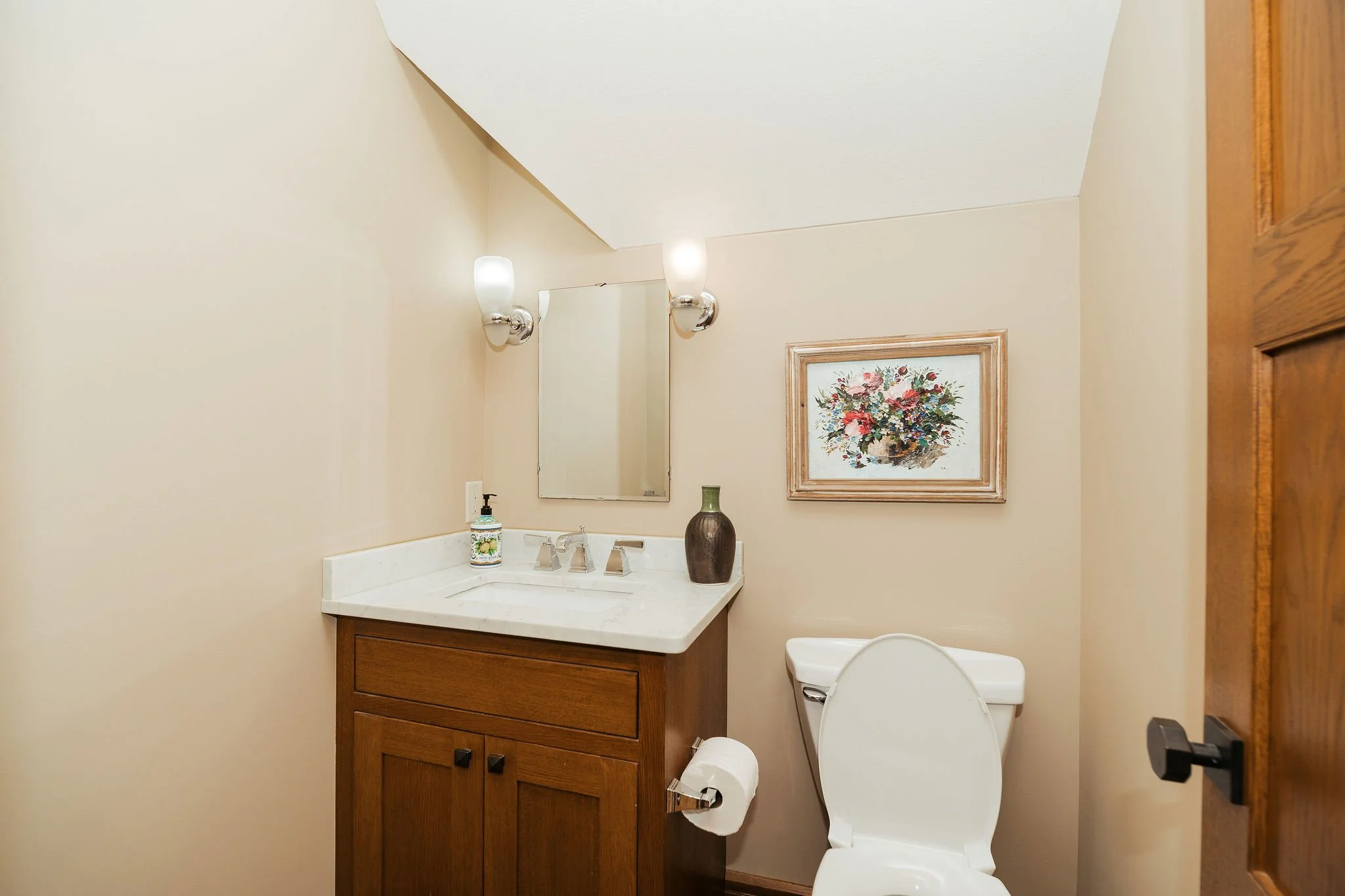 A small bathroom with a wooden vanity, a white marble countertop, a rectangular mirror, a wall-mounted light fixture, a soap dispenser, a vase, and a framed floral painting on the wall, with a white toilet next to the vanity.